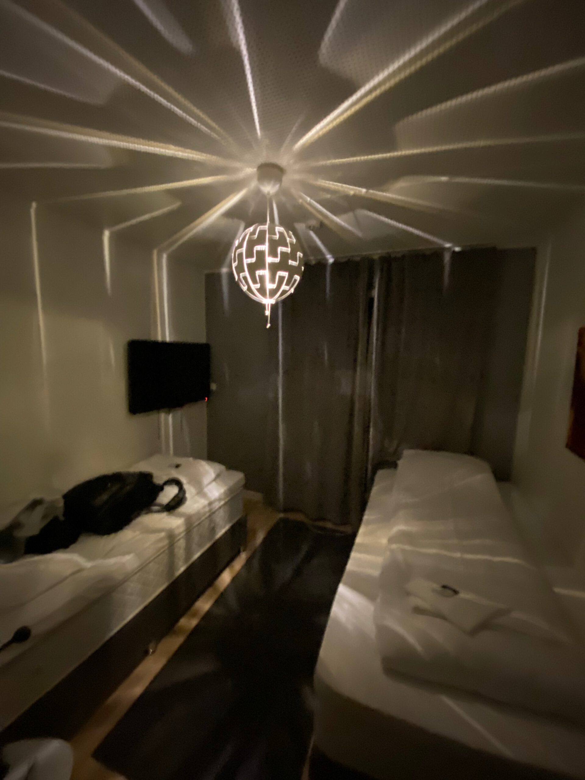Bedroom at Mosjøen Romutleie Sentrum 3 with two single boxspring beds, wall mounted television, rug, and distinctive ceiling lamp creating light patterns, a simple budget accommodation room in Mosjøen
