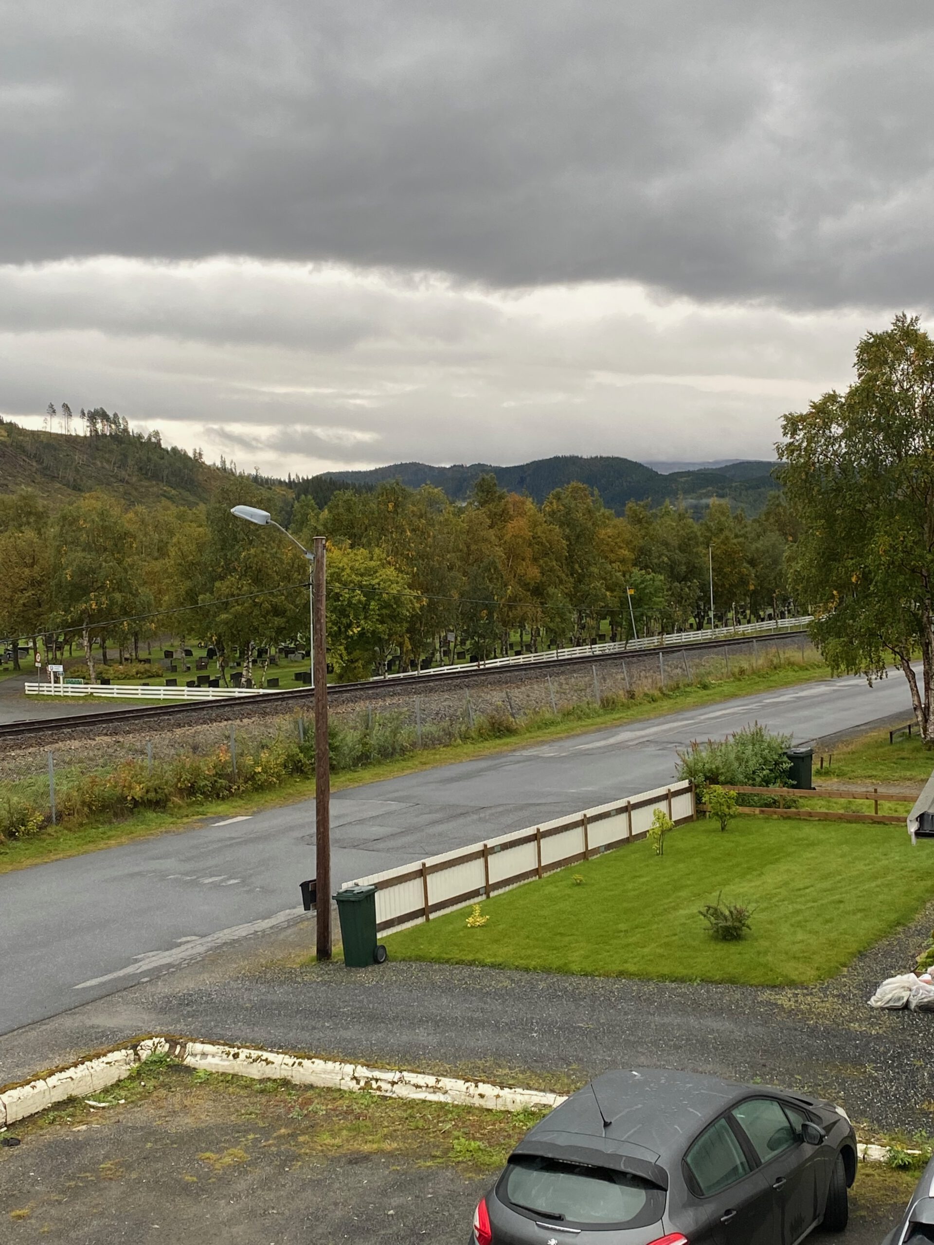 View from Mosjøen Romutleie Sentrum 3 overlooking a quiet street, railway line, green garden, cemetery, and forested hills under an overcast sky, a typical Mosjøen Norway surroundings view from the guesthouse