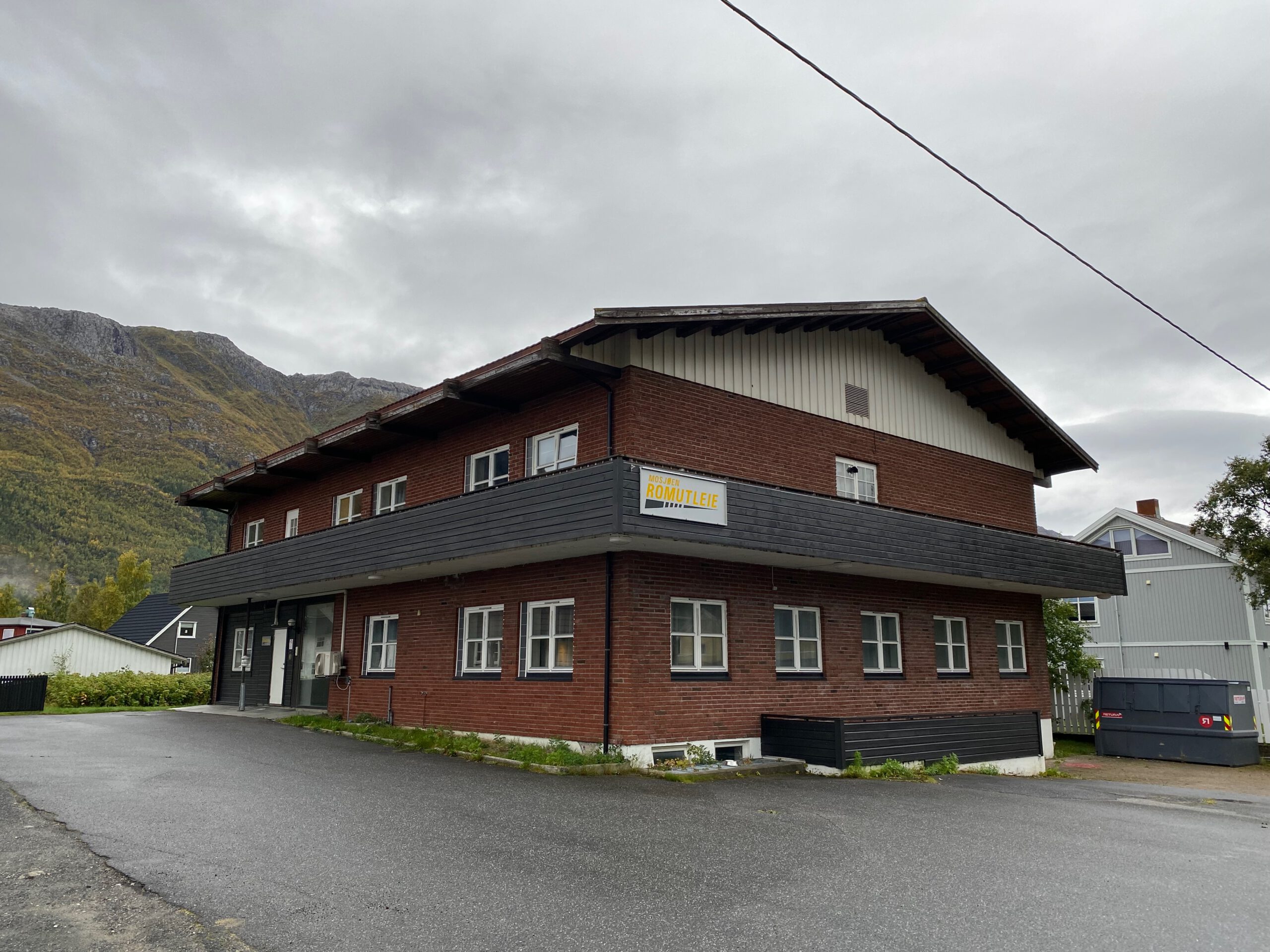 Exterior view of Mosjøen Romutleie Sentrum 3 in Mosjøen, a budget guesthouse with brick facade, balconies, parking area, and surrounding Norwegian mountains under an overcast sky