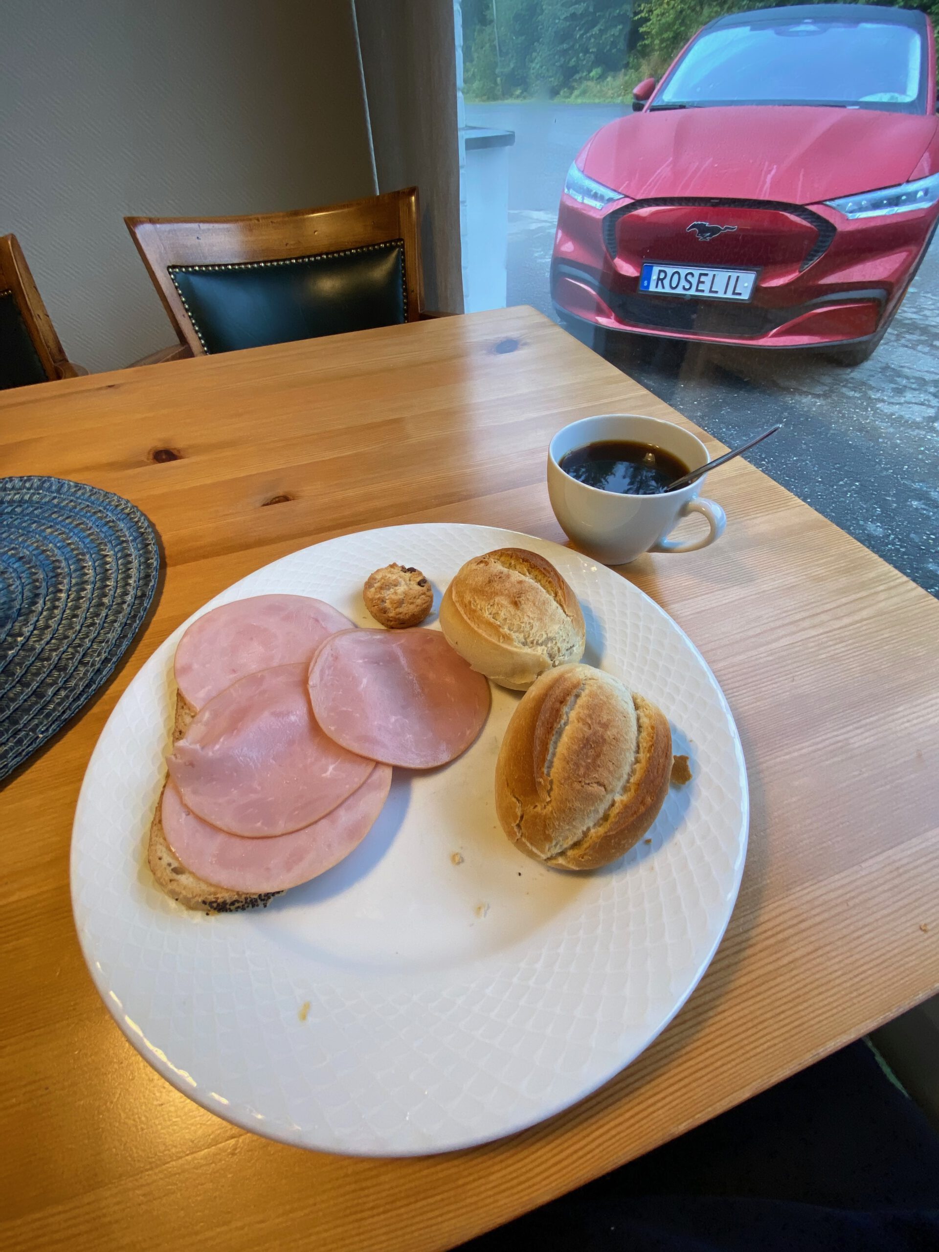 Støren Hotel Norway review breakfast plate with sliced ham on bread rolls, fresh buns and black coffee inside the dining room in Trøndelag, with a red Ford Mustang Mach E parked outside the window