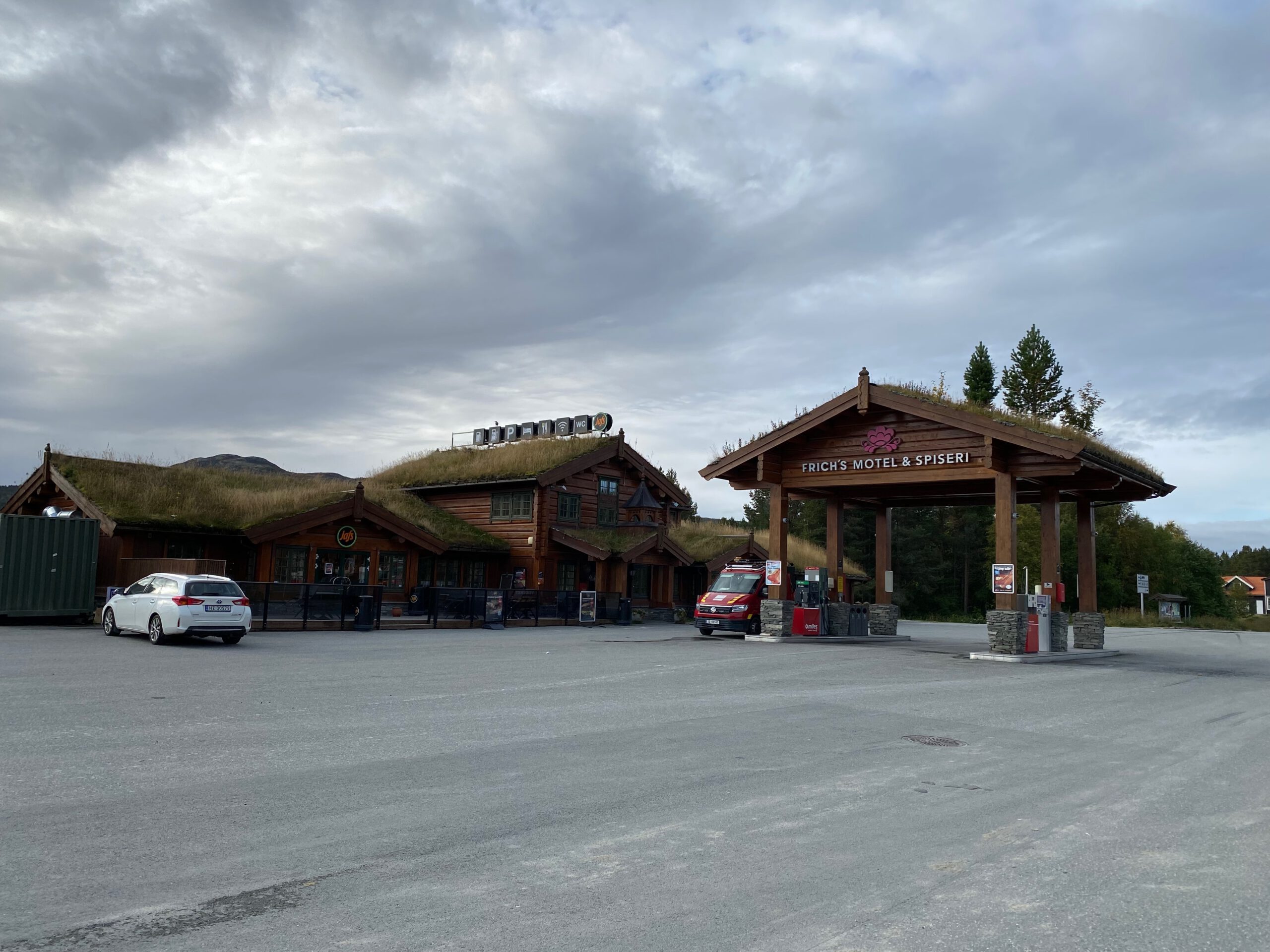 Wide exterior view of Oppdalsporten Fagerhaug roadside stop in Norway, including Frich’s Motel & Spiseri log buildings with traditional grass roofs, Circle K Automat fuel station, parking area with cars and camper vehicles, located along the E6 near Oppdal.