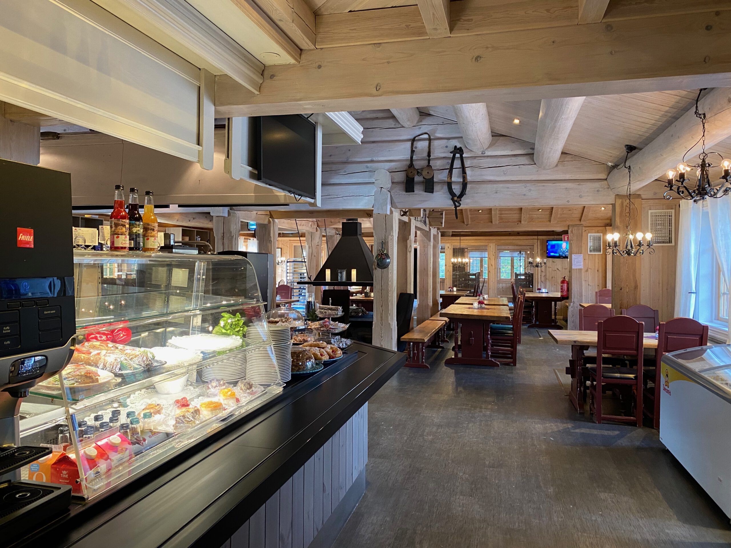 Interior of Oppdalsporten Fagerhaug restaurant inside Frich’s Motel & Spiseri, showing a Friele coffee machine, pastry display with Norwegian baked goods, wooden log cabin interior, fireplace, chandelier lighting, dining tables and cozy Scandinavian mountain lodge atmosphere.