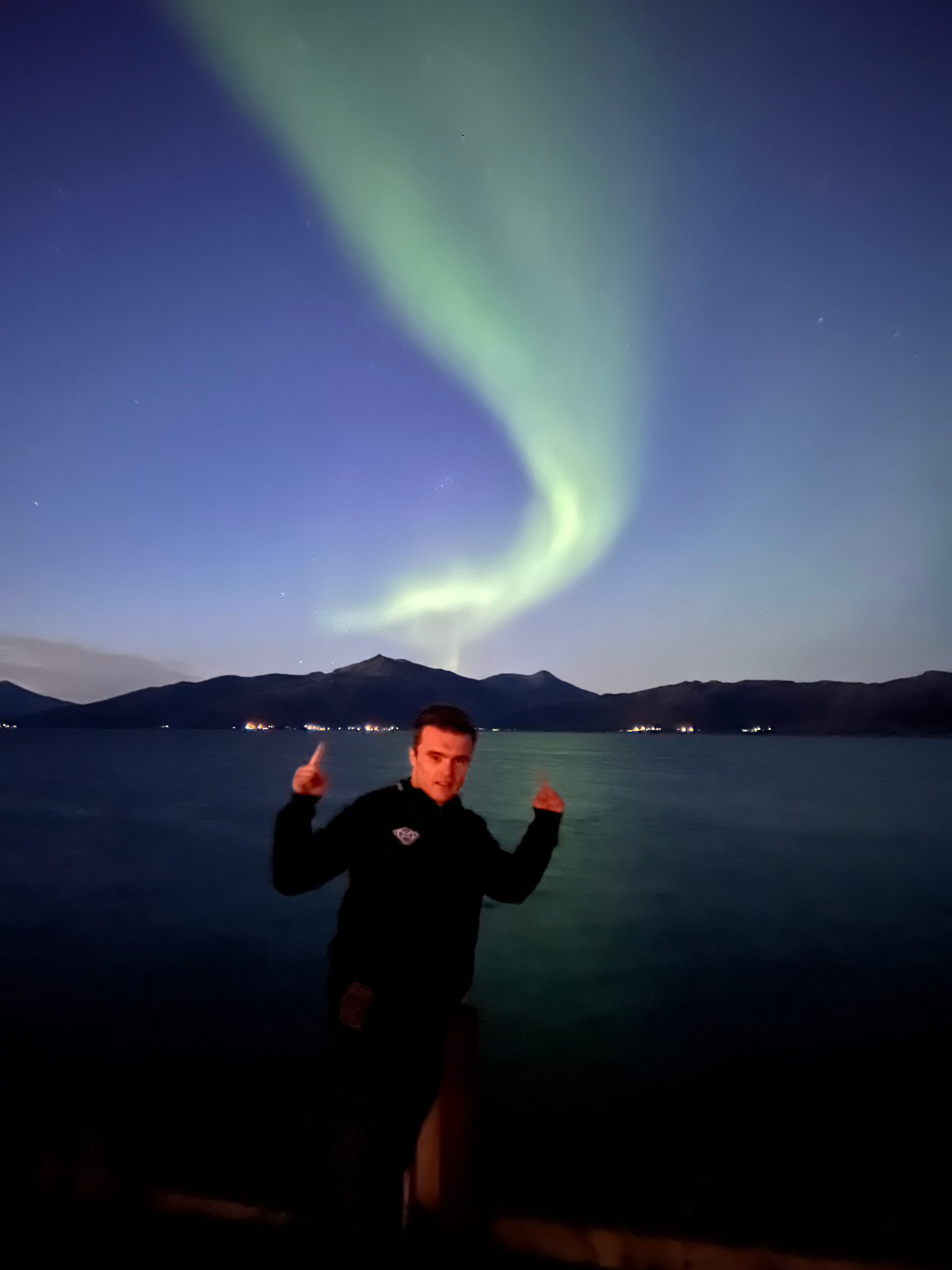 Hjortholmen Northern Lights Spot with a sweeping aurora borealis above the coastline of Lødingen, photographed from the pier with mountains and calm water in the background with Peter Smit on front