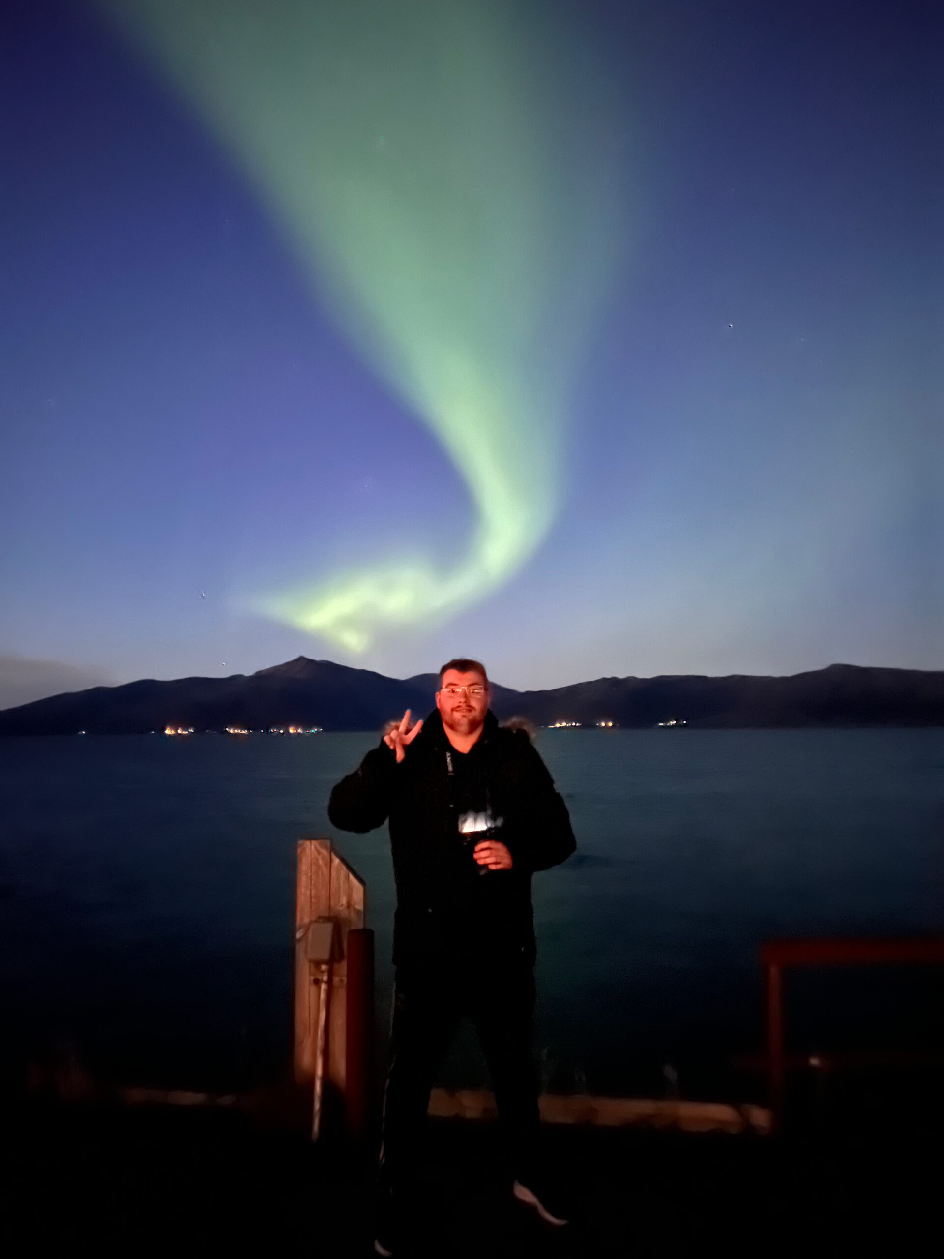 Hjortholmen Northern Lights Spot showing a bright green aurora above the sea near Lødingen, with Timo van den Berg / Timeuw standing on a wooden pier under the Arctic night sky