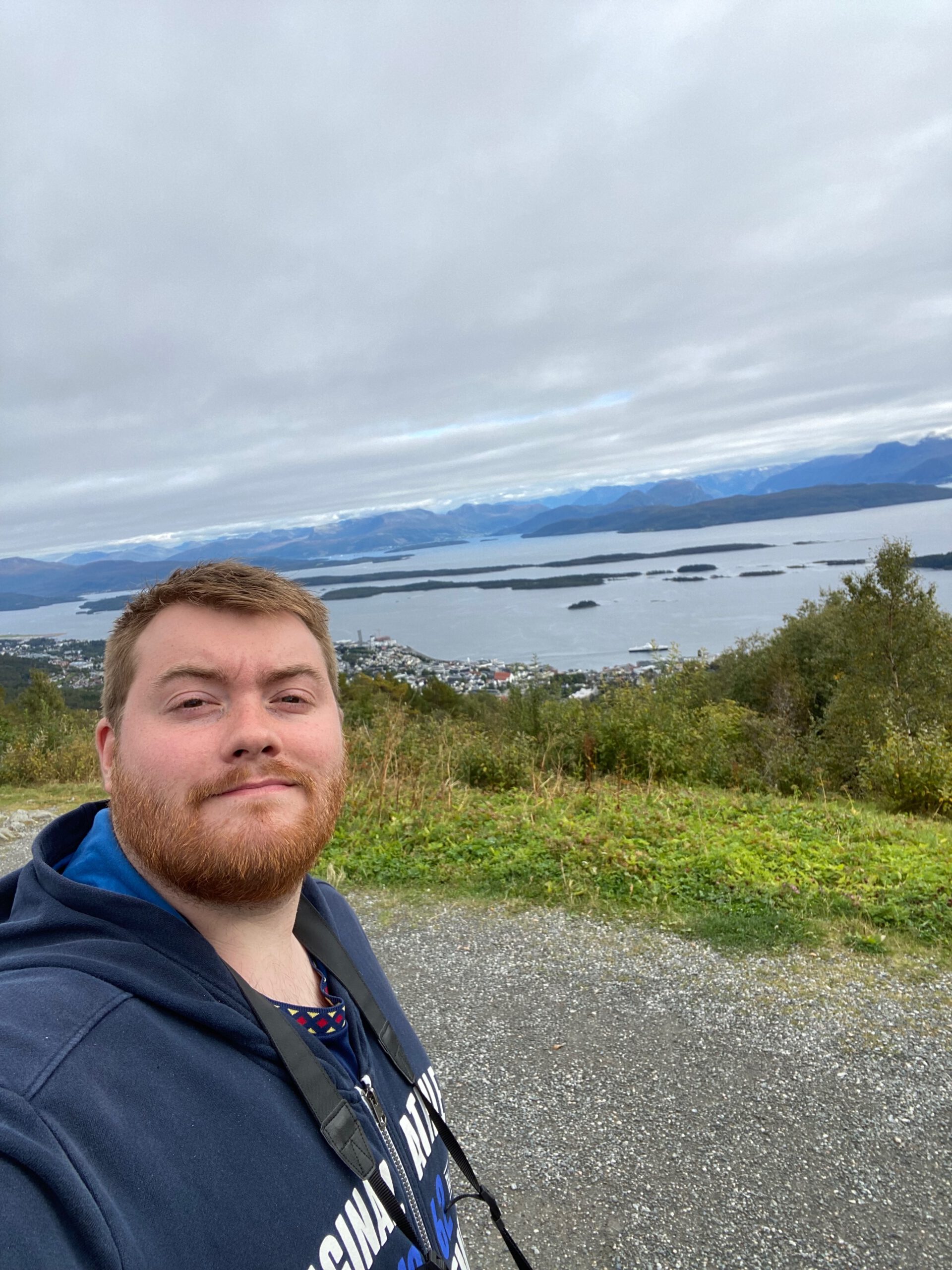 Vardestua Restaurant Molde view selfie at 407 meters above sea level, panoramic fjord and island landscape behind visitor overlooking Molde Norway under dramatic cloudy sky.