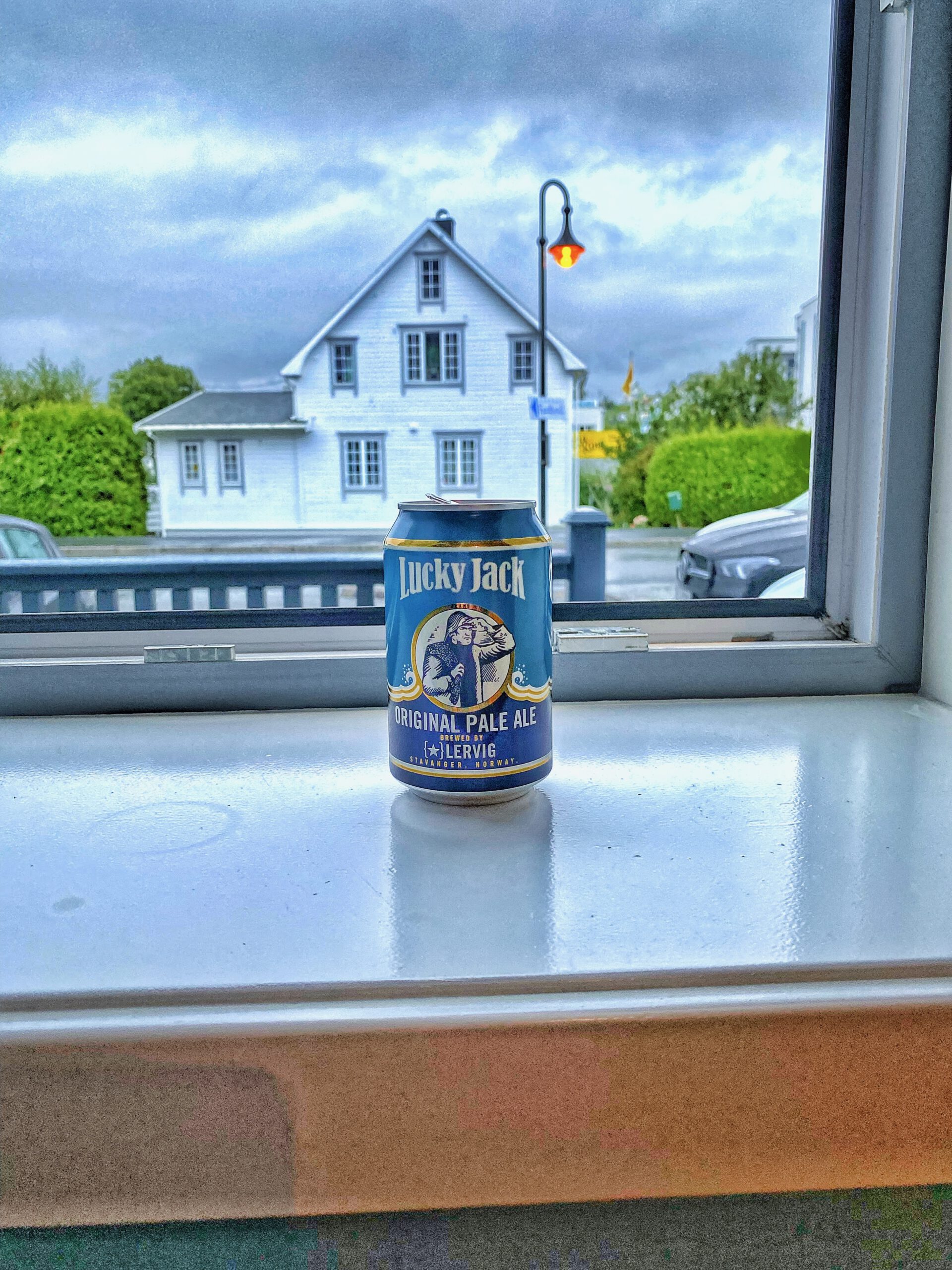Lucky Jack Original Pale Ale beer on windowsill inside Vestnes Fjordhotel with Norwegian house and rainy fjord village street in background