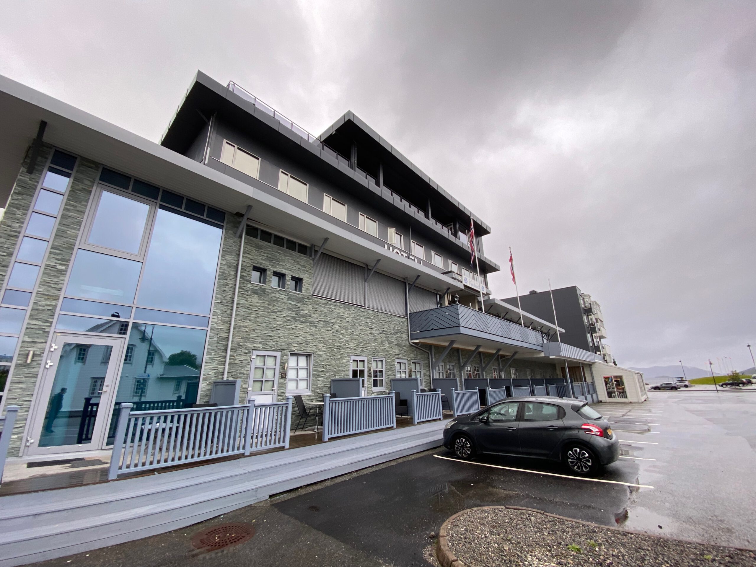 Vestnes Fjordhotel exterior in Vestnes Norway with modern stone facade, balconies, Norwegian flags, and parked car under cloudy fjord sky