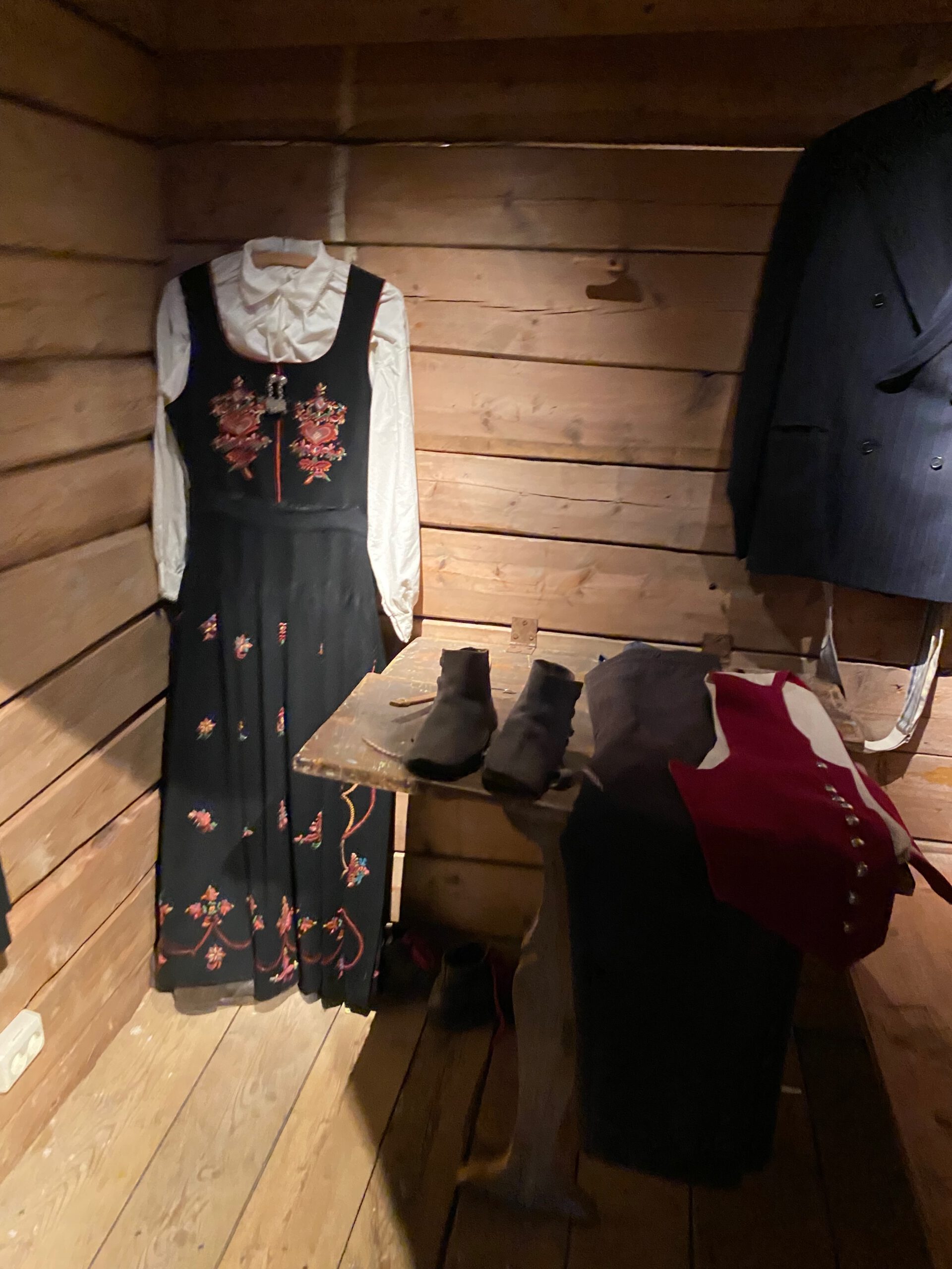 Traditional Norwegian folk costumes exhibited at Norsk Fjordsenter Geiranger featuring embroidered bunad dress, vest and boots inside wooden cabin setting, part of the UNESCO World Heritage Geirangerfjord cultural display