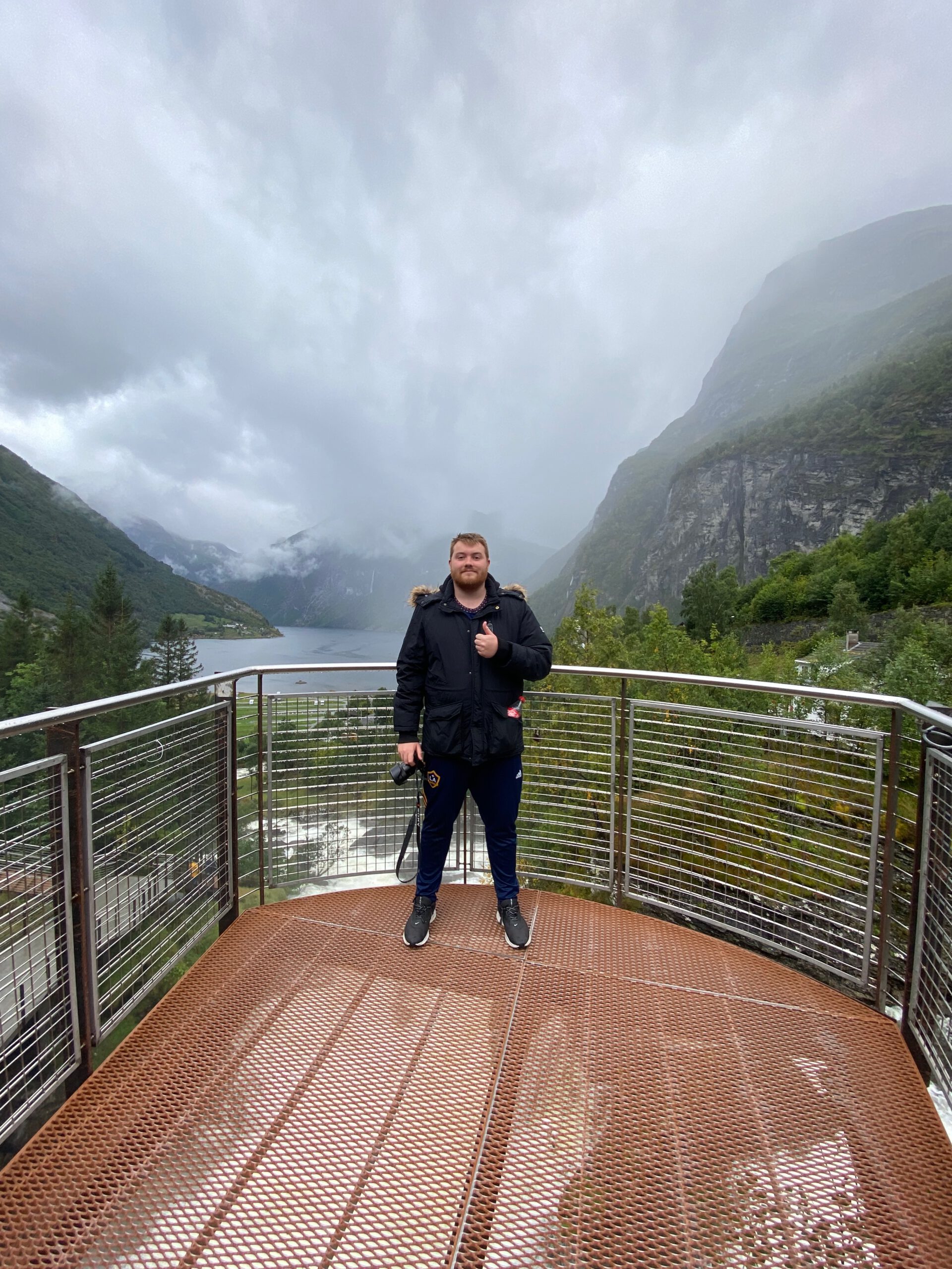 Timo van den Berg aka Timeuw standing on viewing platform at Norsk Fjordsenter Geiranger overlooking the UNESCO World Heritage Geirangerfjord surrounded by steep mountains and misty fjord landscape