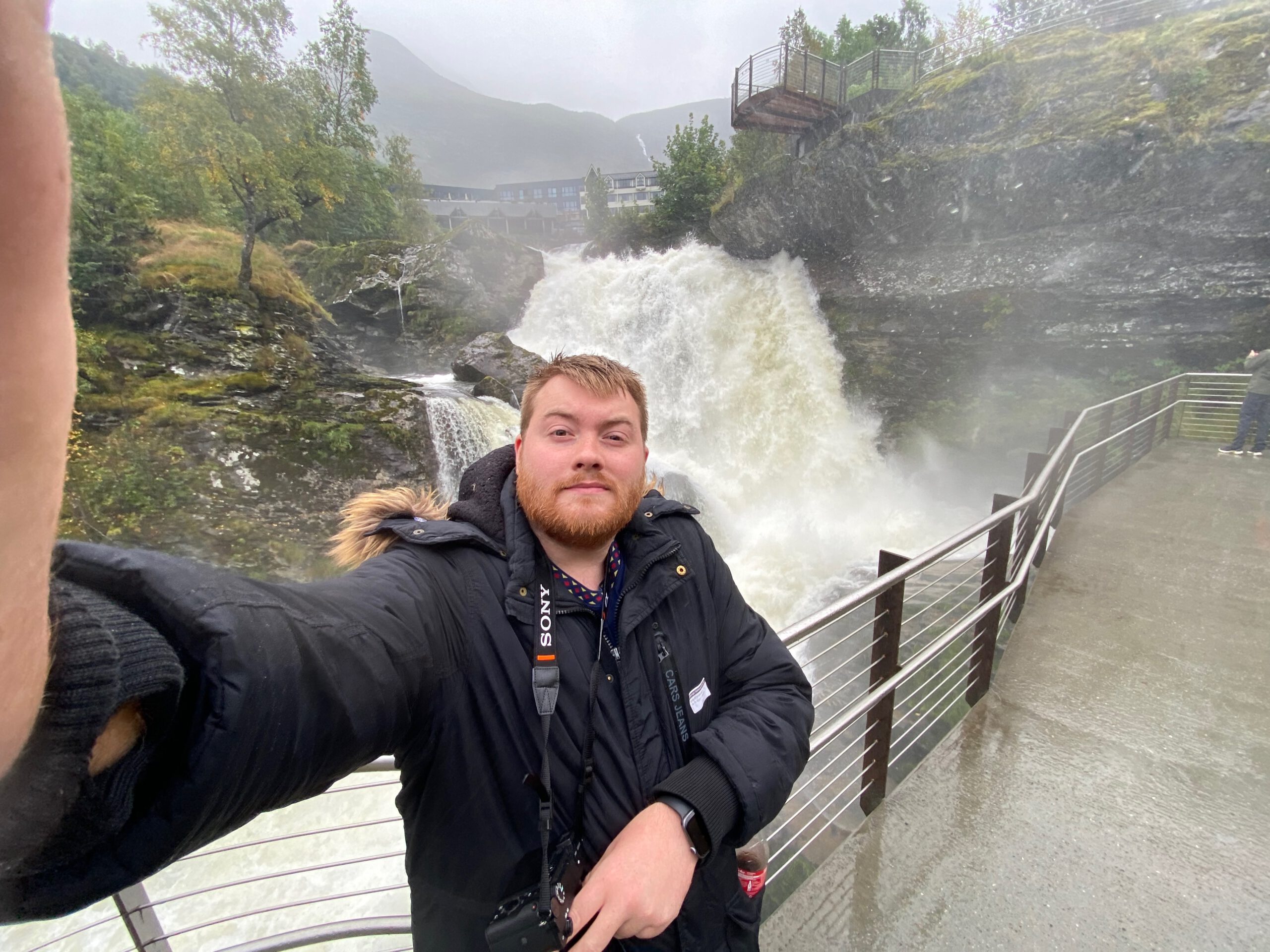 Timo van den Berg aka Timeuw taking selfie at Norsk Fjordsenter Geiranger with powerful waterfall and misty UNESCO World Heritage Geirangerfjord landscape in Norway