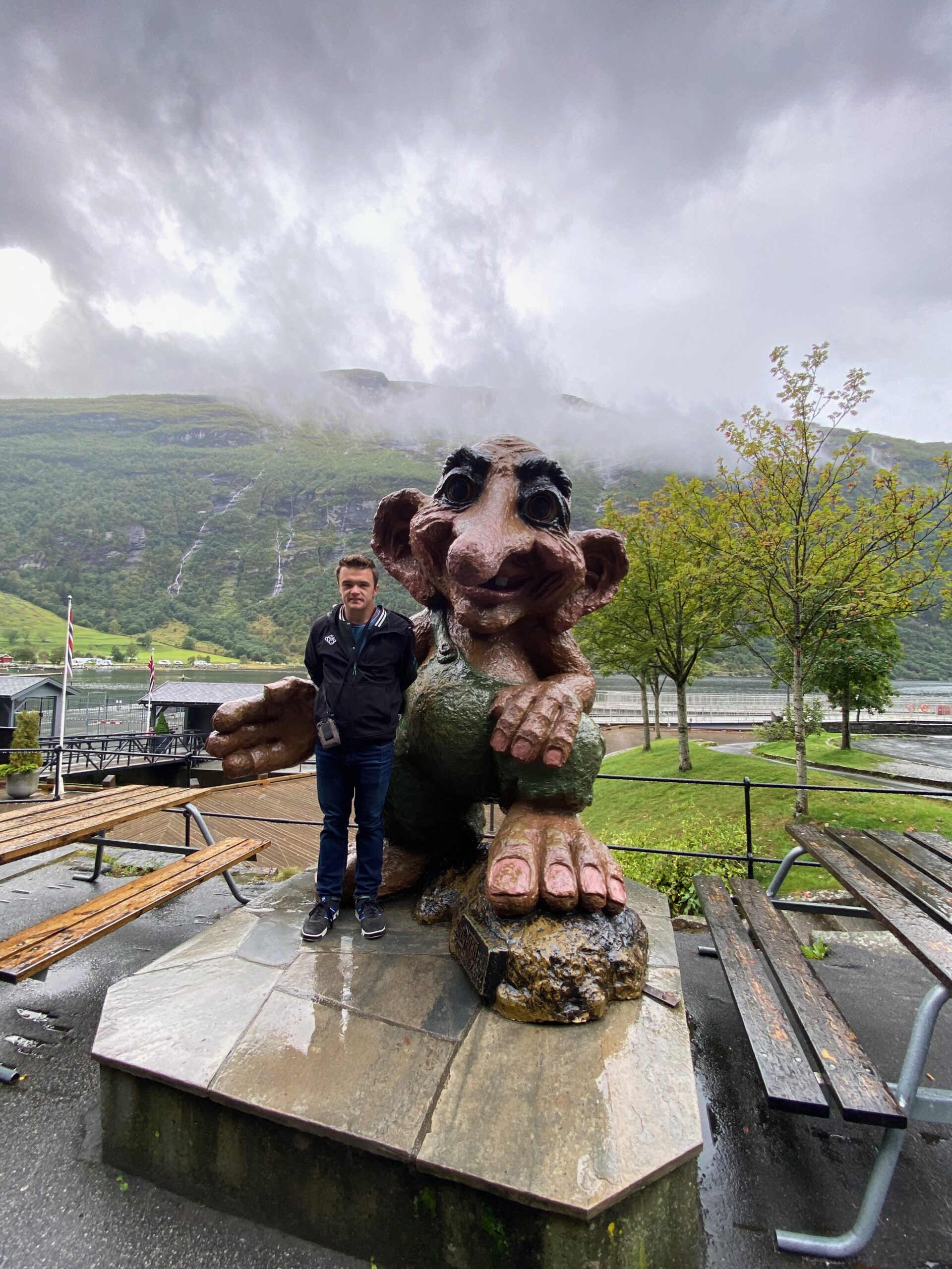 Peter Smit standing next to the troll statue in Geiranger near the harbor before the Geiranger Fjord and Waterfalls Sightseeing Boat Trip with waterfalls and fjord landscape behind