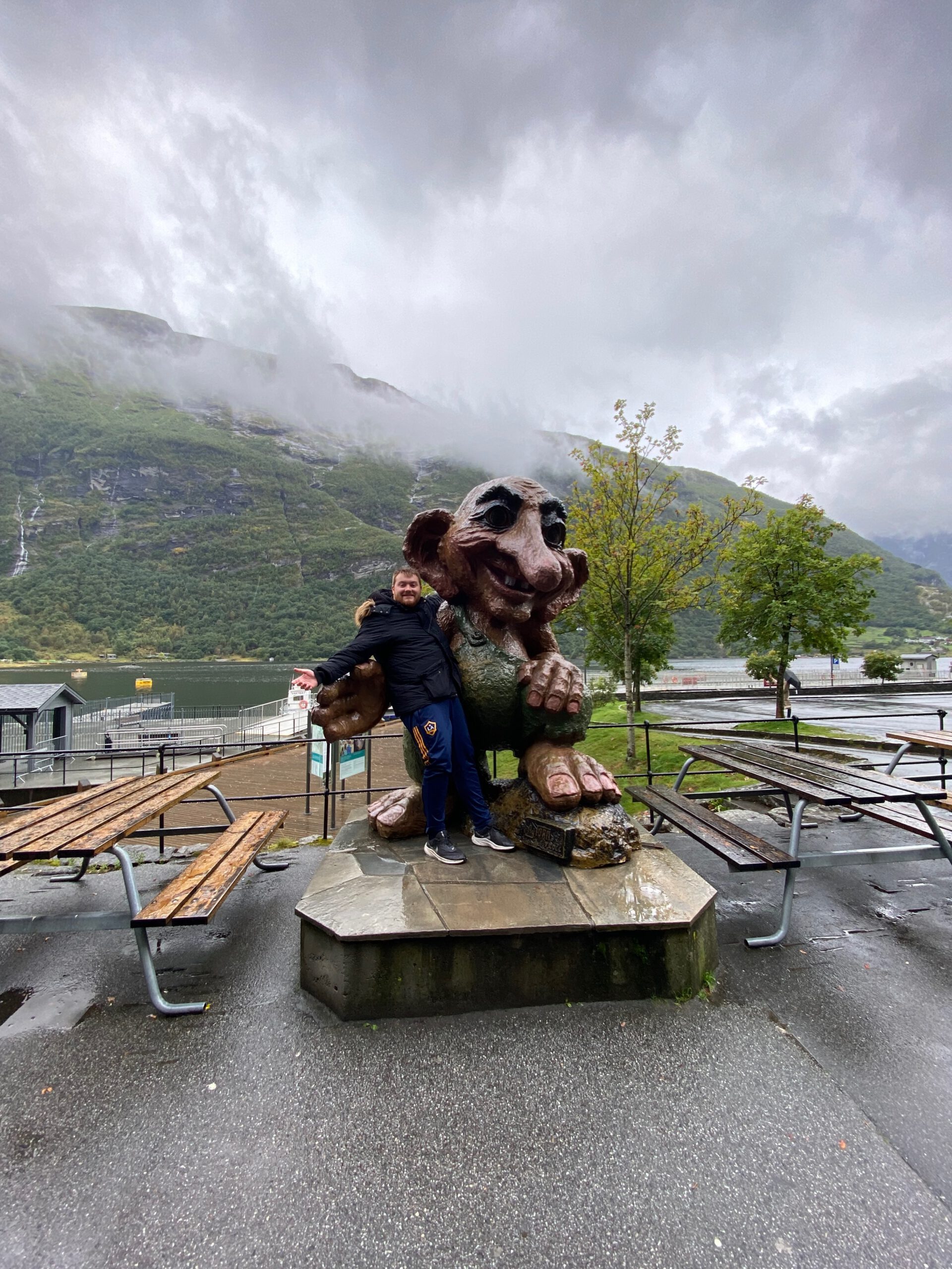Timo van den Berg Timeuw posing with the troll statue in Geiranger village during the Geiranger Fjord and Waterfalls Sightseeing Boat Trip with misty mountains and fjord in the background
