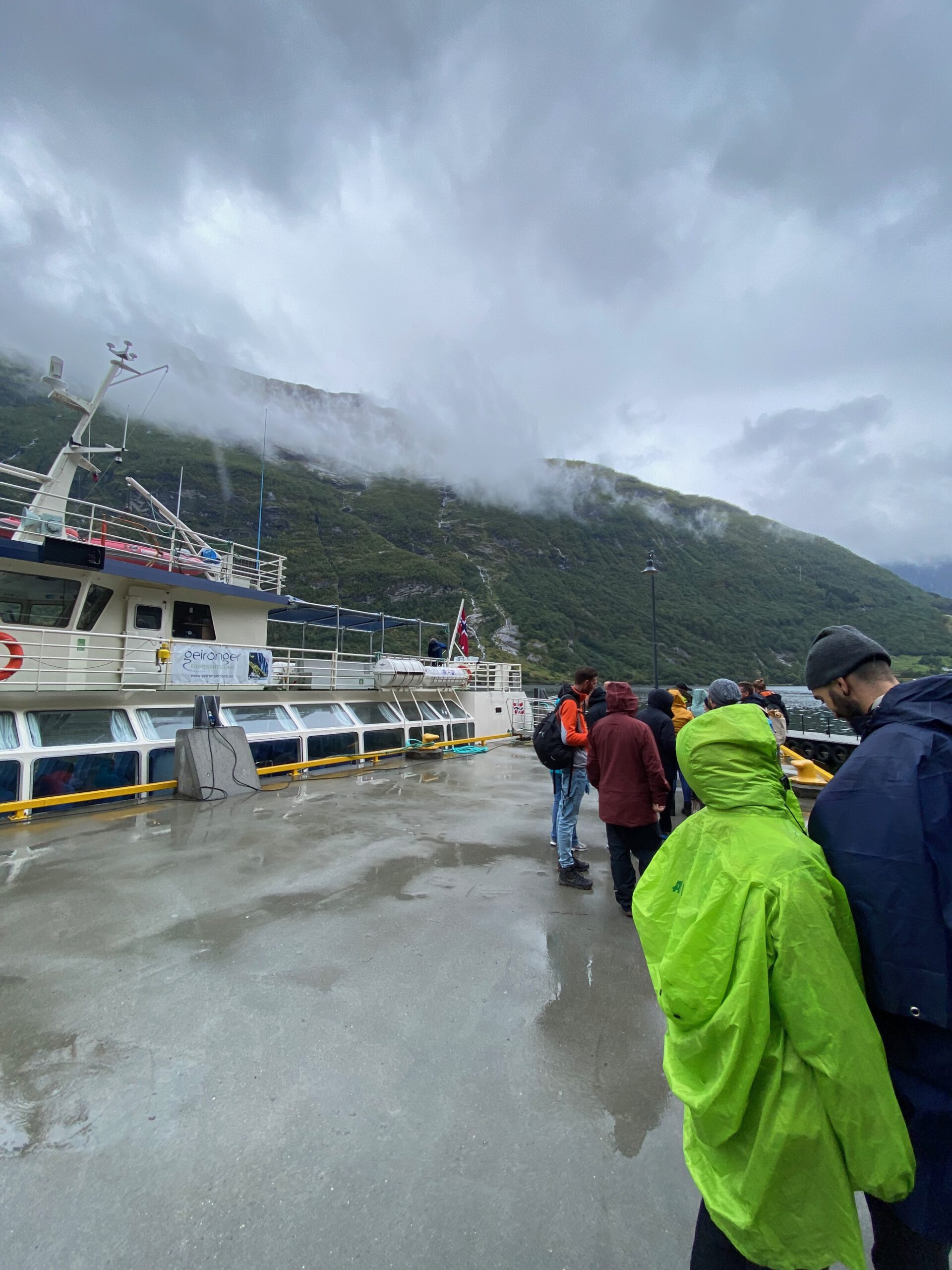 Passengers boarding at Geiranger harbor during the Geiranger Fjord and Waterfalls Sightseeing Boat Trip with rainy weather and misty mountains in the background