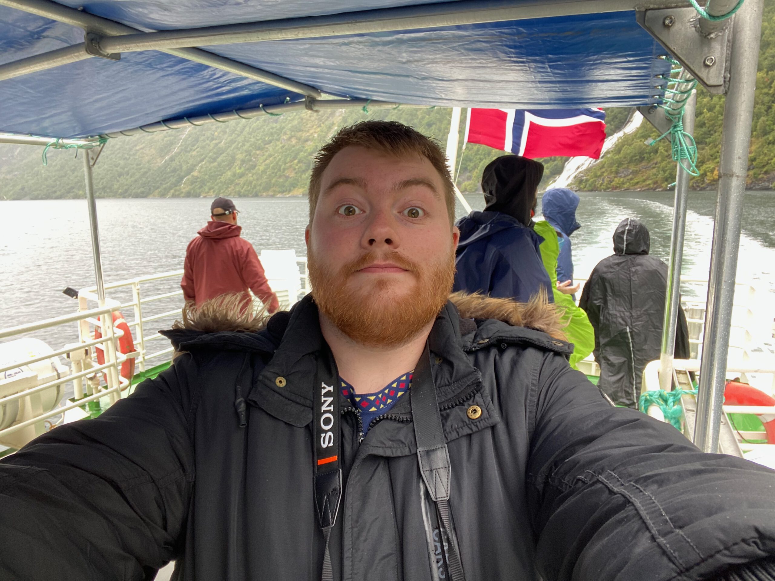 Timo van den Berg Timeuw during the Geiranger Fjord and Waterfalls Sightseeing Boat Trip standing on the outdoor deck with Norwegian flag and dramatic Geirangerfjord cliffs in the background