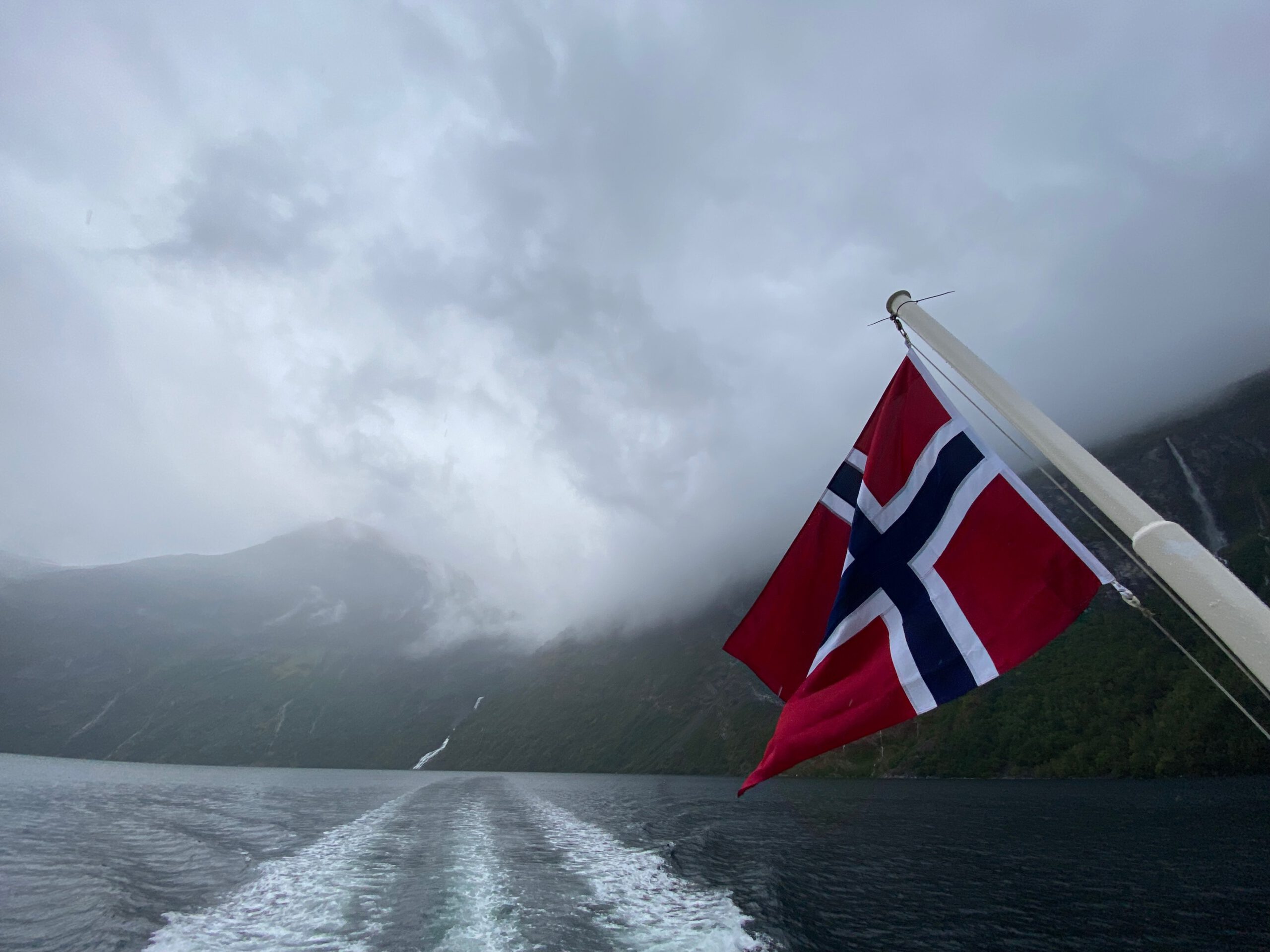 Norwegian flag waving during the Geiranger Fjord and Waterfalls Sightseeing Boat Trip with misty mountains and dramatic Geirangerfjord scenery in the background