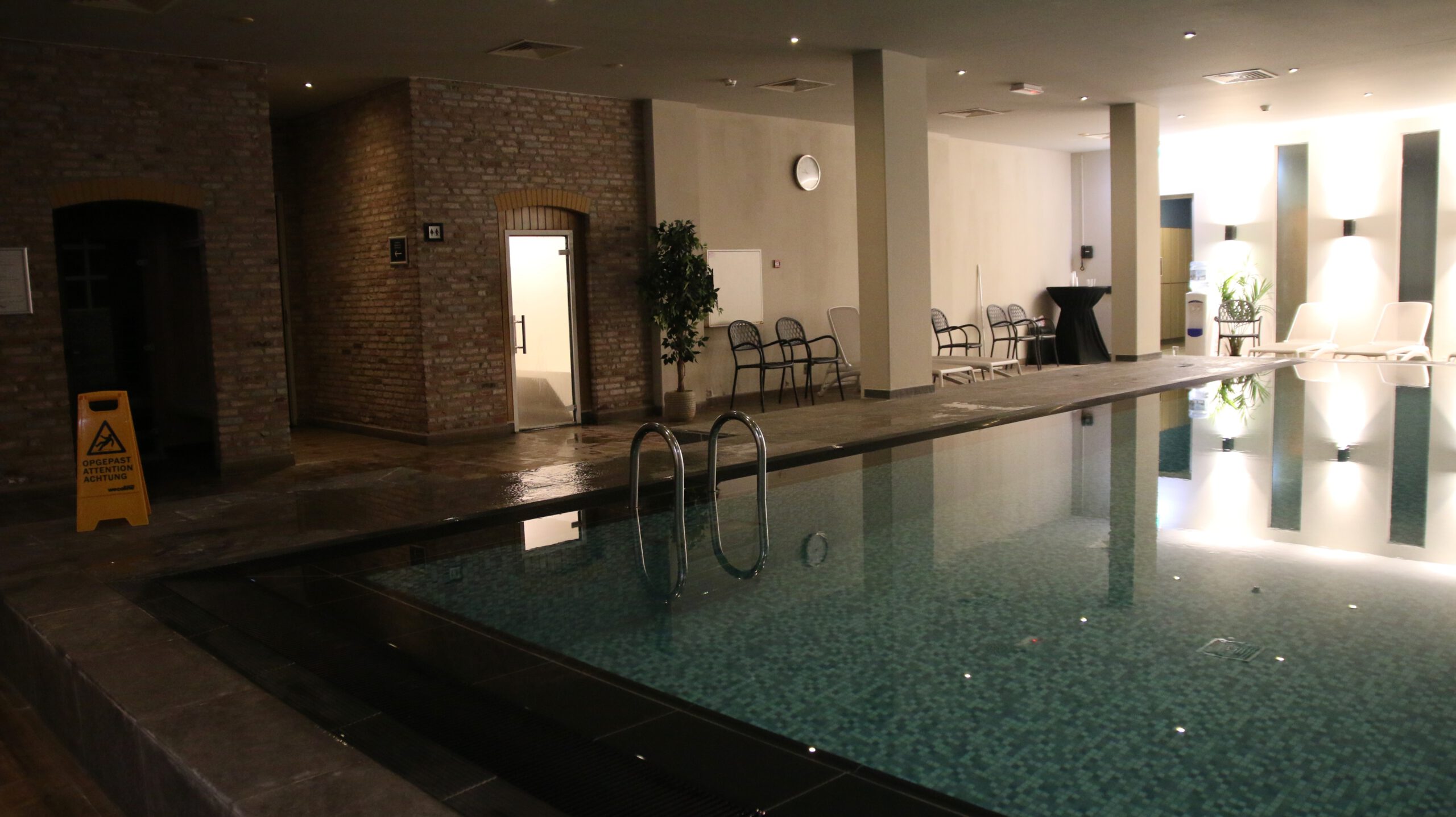 Grand Hotel Ter Duin review indoor spa facilities in Burgh Haamstede featuring a heated swimming pool with ladder access, mosaic tile floor, brick sauna entrance, lounge chairs, changing area, and wellness seating area