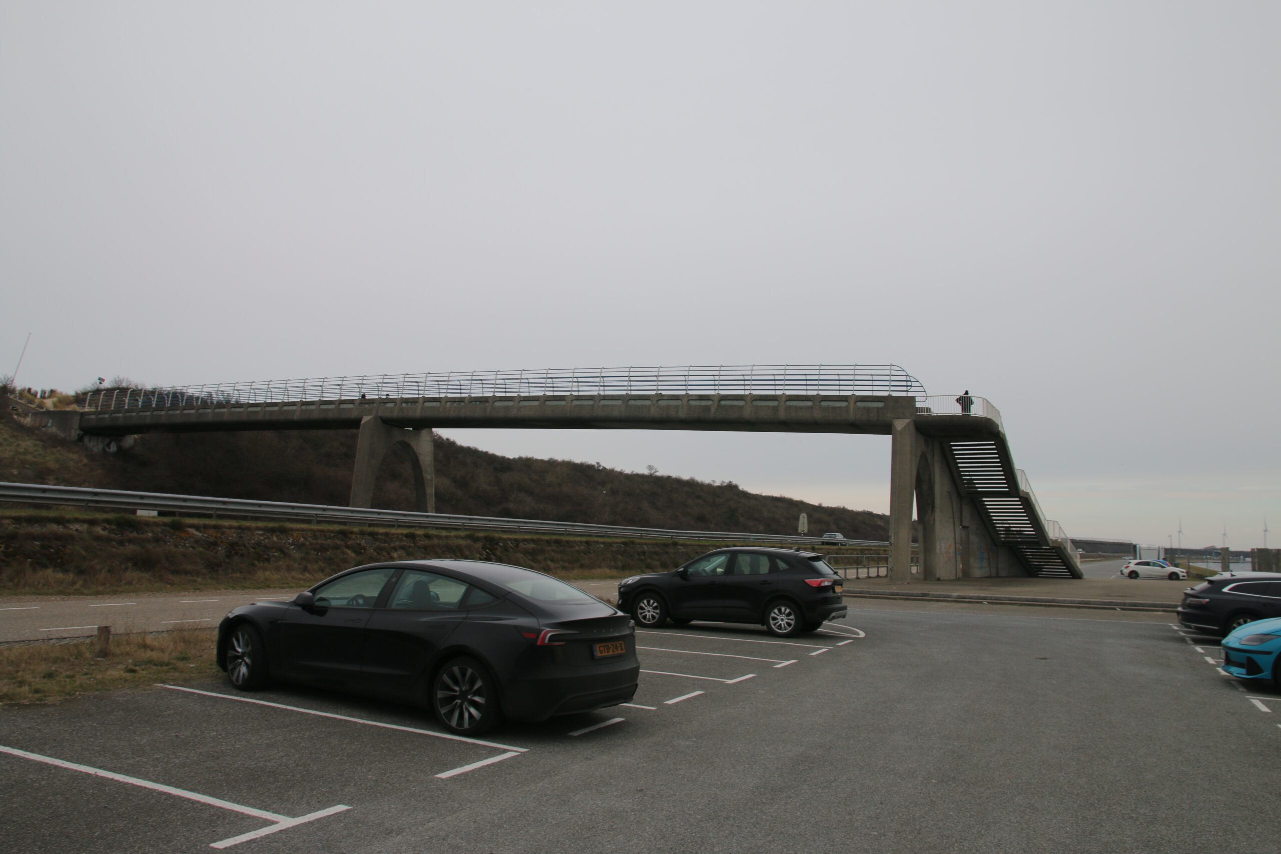 Strandrestaurant De Dam access route via elevated pedestrian bridge and concrete staircase over the main coastal road in Vrouwenpolder, parking lot with multiple cars including a black sedan, dunes and wind turbines visible in the distance, gray cloudy sky and open coastal landscape surrounding the beach area.