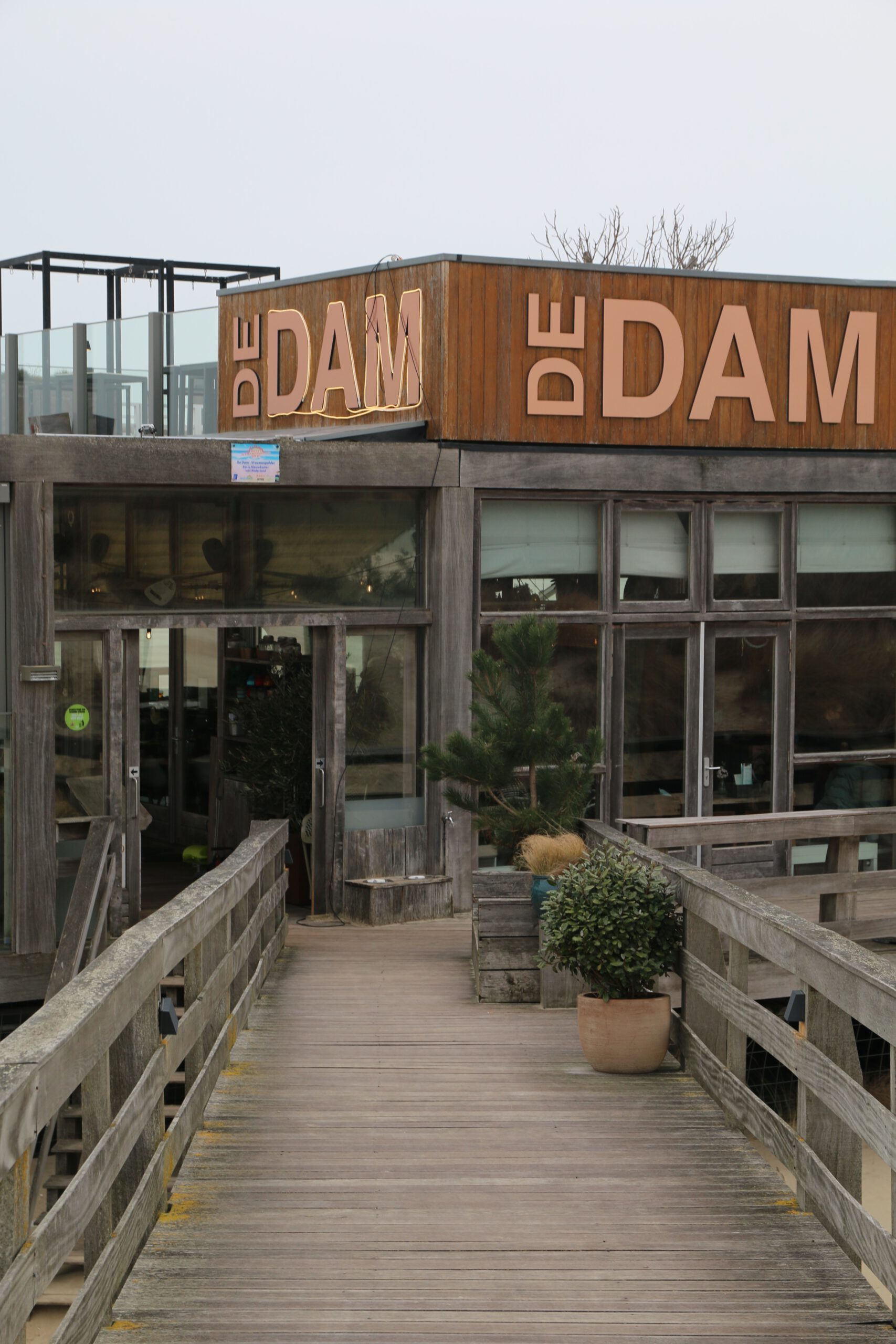 Strandrestaurant De Dam review entrance view with wooden walkway leading to the beach pavilion facade and De Dam signage above the glass windows.