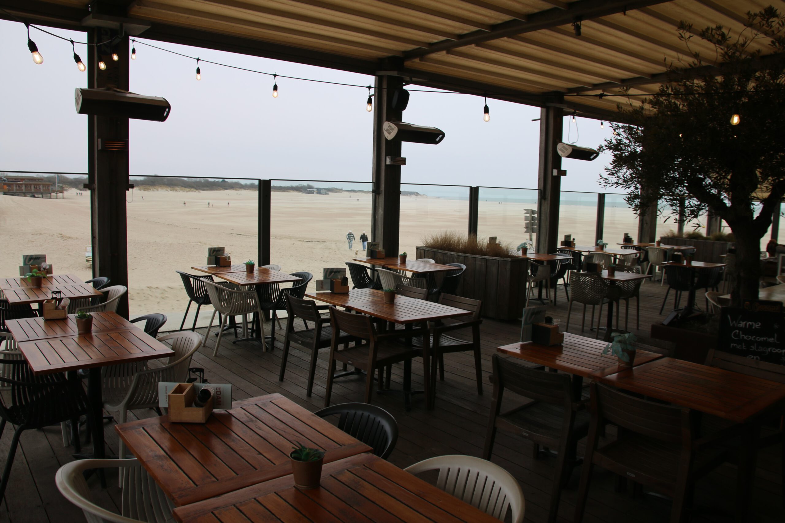 Strandrestaurant De Dam covered terrace interior with wooden tables and mixed chairs, hanging string lights under the roof, outdoor heaters mounted on black pillars, glass wind panels facing the sandy beach and North Sea, few people walking in the distance, large potted tree inside, cozy beach pavilion atmosphere in Vrouwenpolder.