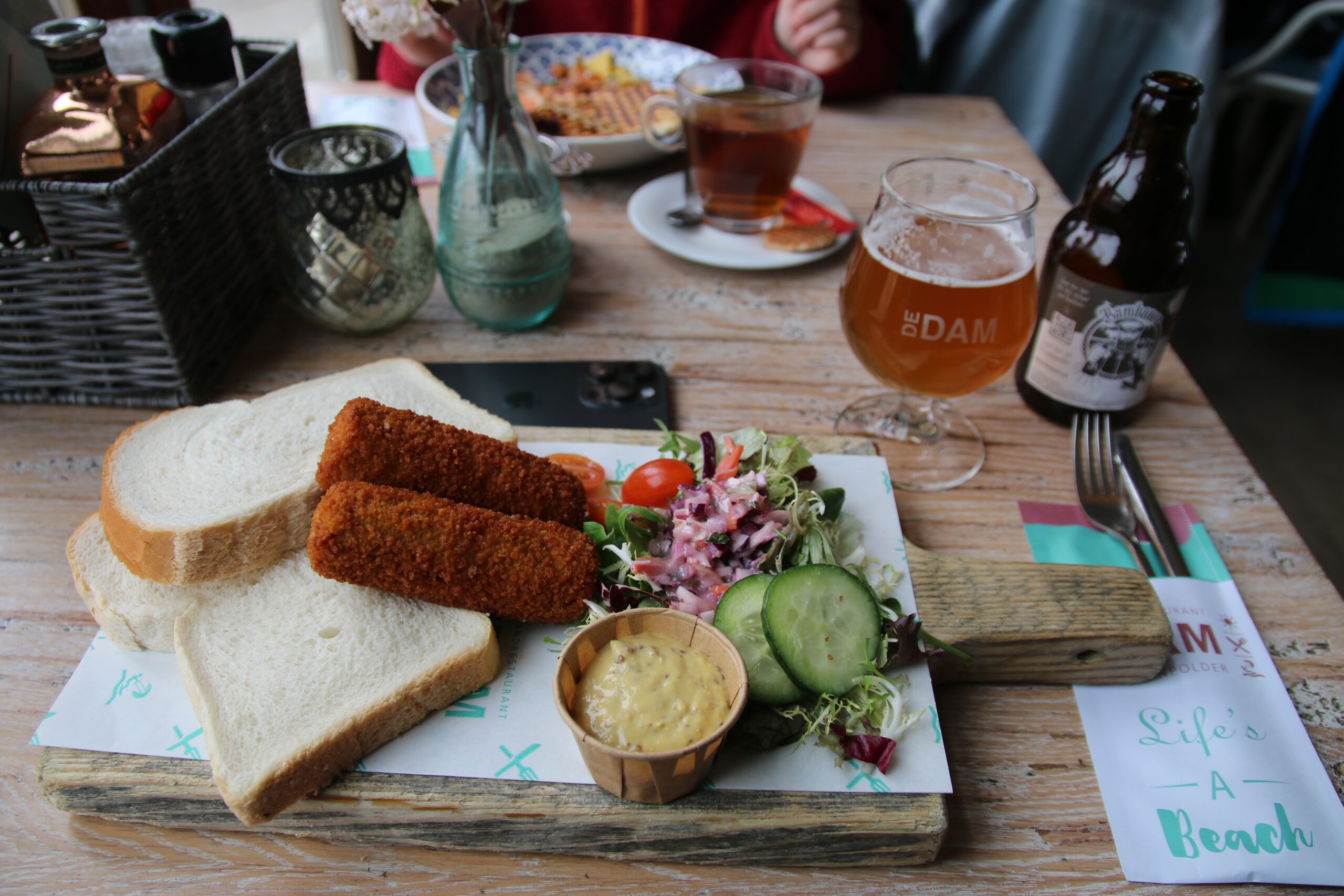 Strandrestaurant De Dam lunch plate with two crispy beef croquettes served on white bread, small wooden cup of mustard, fresh mixed salad with cucumber and cherry tomato, rustic wooden serving board on a beach style table, glass of Bambaver local beer with De Dam logo, beer bottle next to the glass, tea served in the background and wooden interior atmosphere inside the beach pavilion in Vrouwenpolder.
