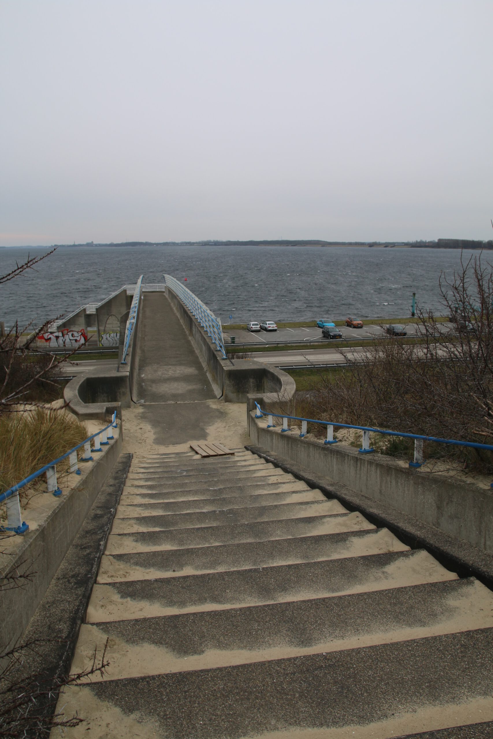 Strandrestaurant De Dam access route via concrete staircase leading down from the dunes to an elevated pedestrian bridge crossing the main coastal road in Vrouwenpolder, Veerse Meer visible in the background, parked cars along the waterfront, gray cloudy sky and sandy dune landscape surrounding the beach entrance area.