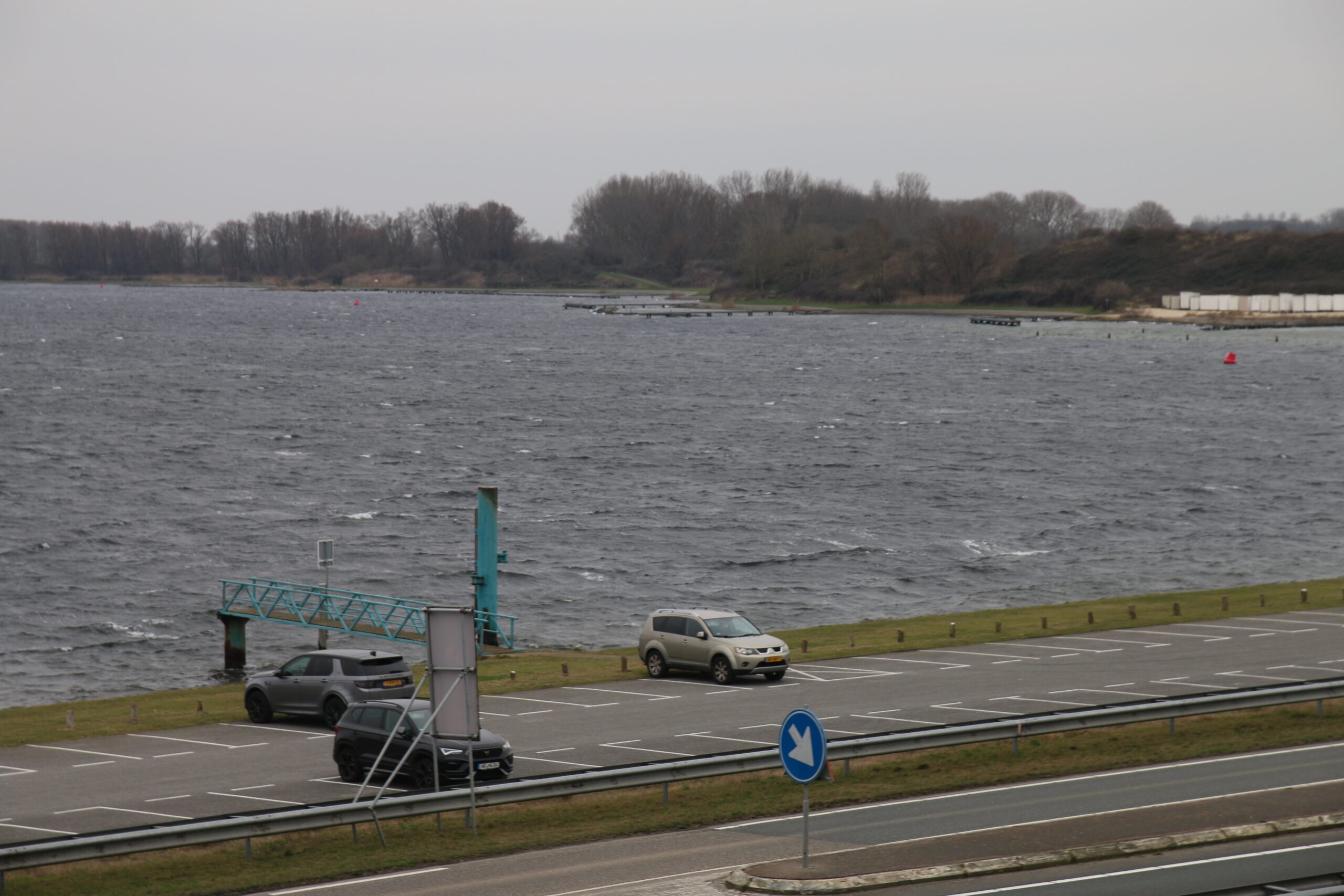 Strandrestaurant De Dam parking option near the Veerse Meer in Vrouwenpolder with several cars parked along the water, small dock structure extending into the lake, windy gray sky, roadside guardrail in the foreground and tree line across the water, showing one of the accessible parking locations close to the beach restaurant.