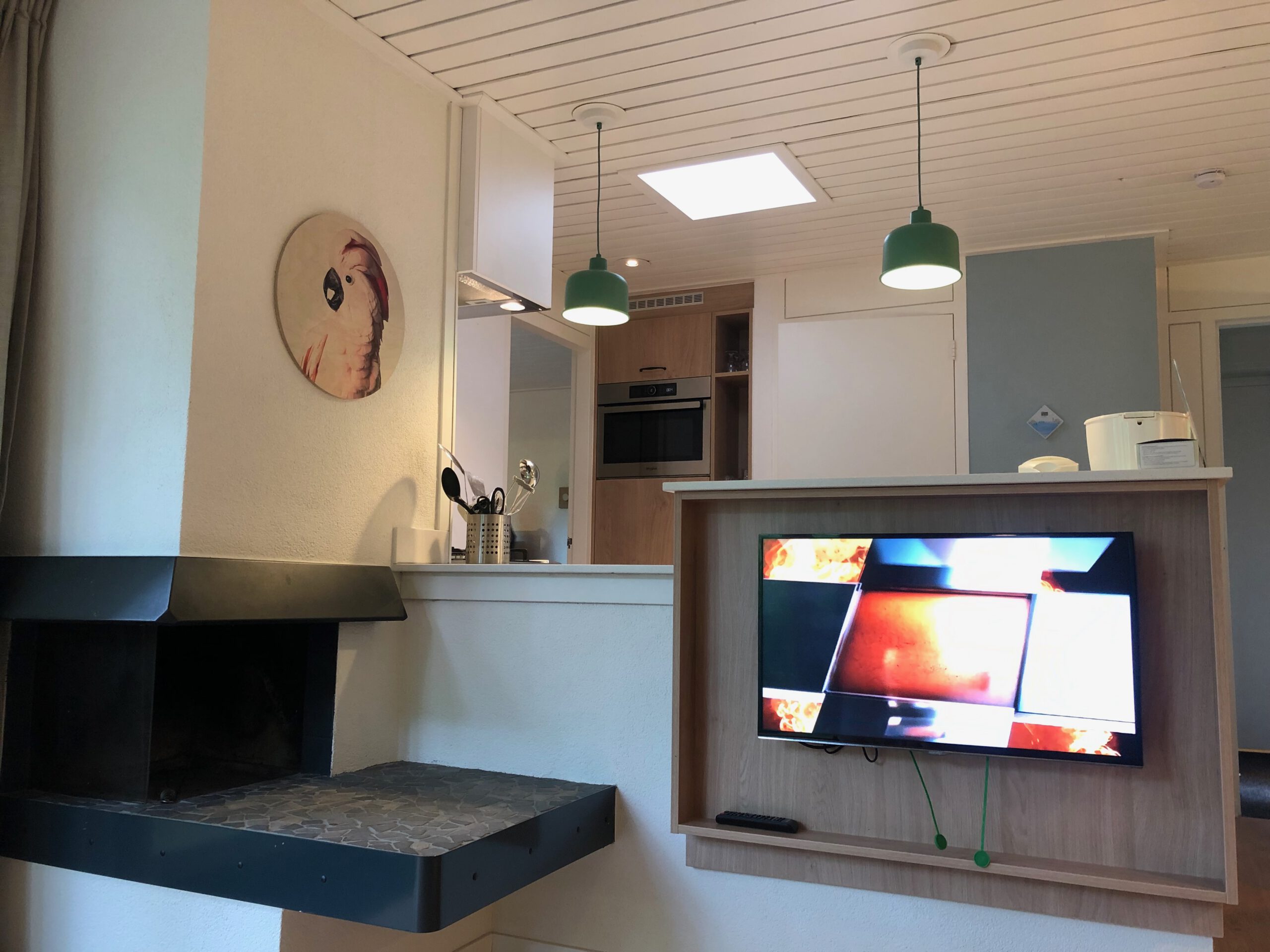 Living area of Center Parcs Het Heijderbos 4 pers. Premium HB1922 cottage 965 showing mounted flatscreen TV, open kitchen layout, green pendant lights and fireplace feature.