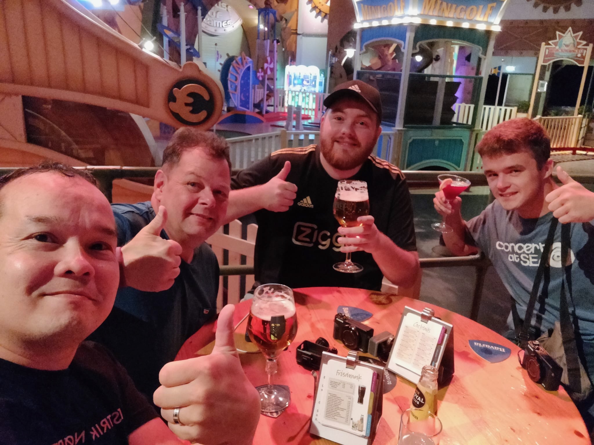 Center Parcs De Huttenheugte Day Visit evening drinks at the Action Factory with Chris and Rogier from AMFVLOGS, Peter Smit, and Timo van den Berg, Timeuw, holding beer and cocktails at a wooden table inside the indoor minigolf area in Dalen, Drenthe.