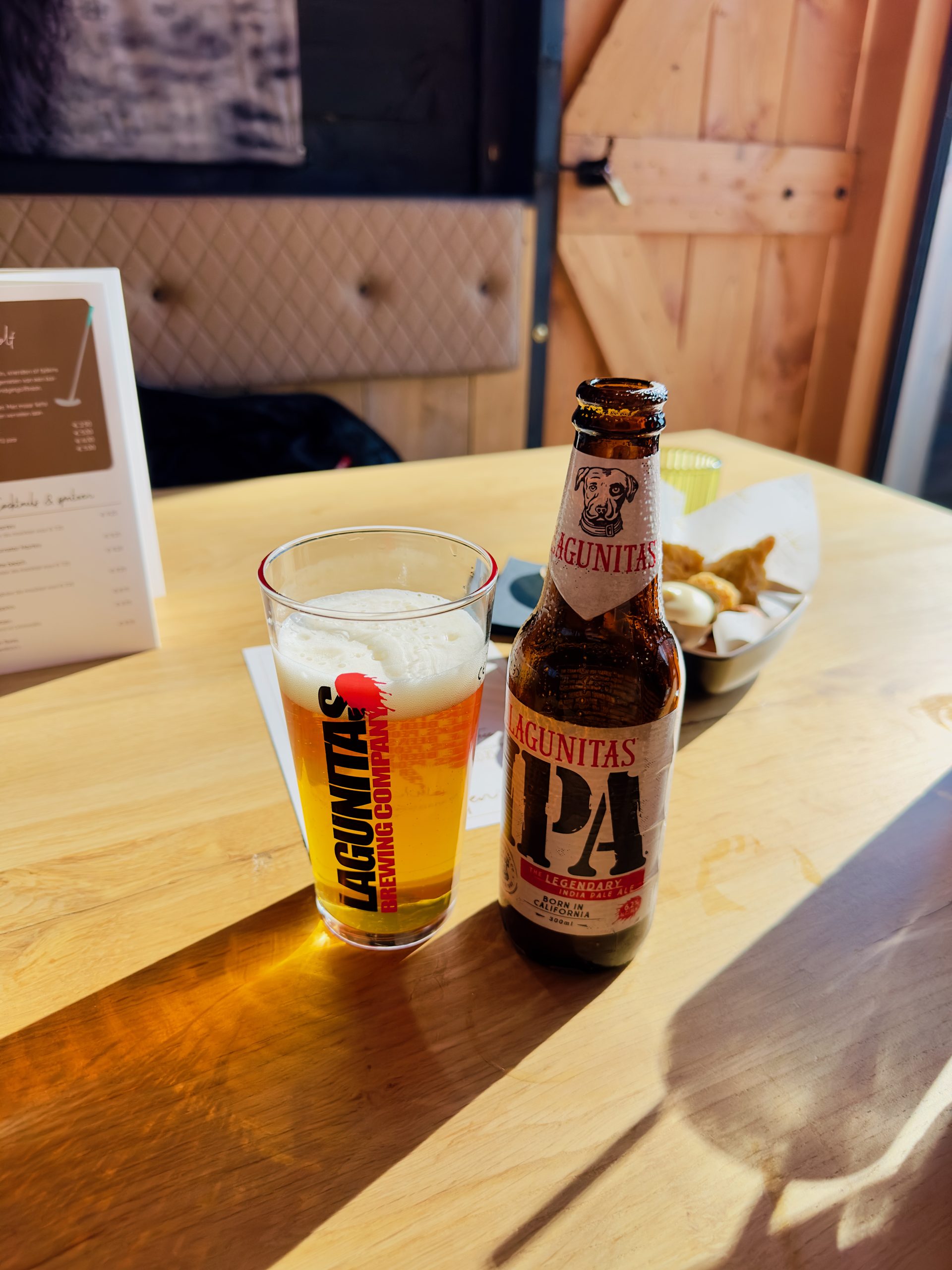 Bij Hen Alphen aan den Rijn, Lagunitas IPA beer with a filled glass and warm bar snacks on a sunny wooden table inside the cozy restaurant.