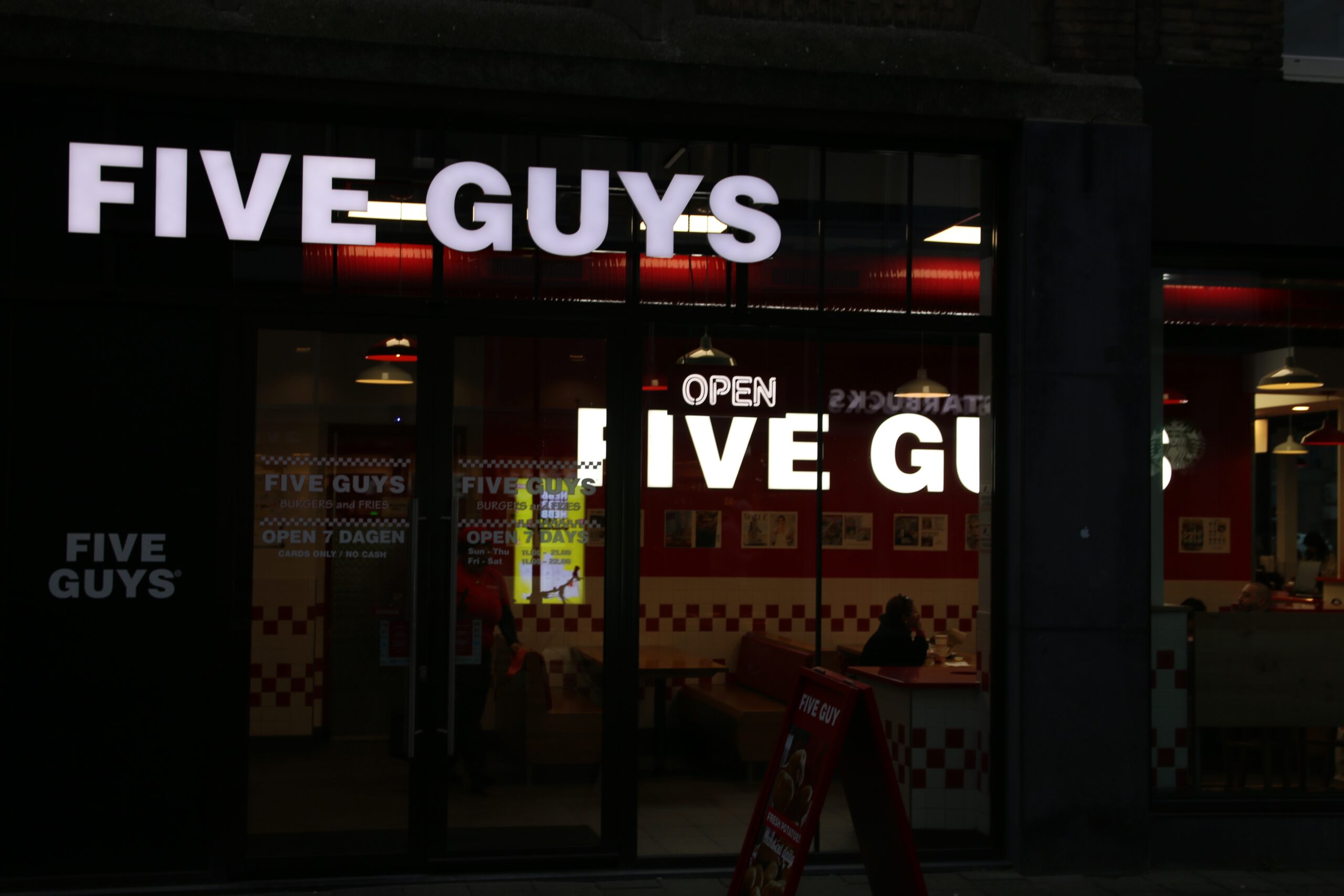 Five Guys Netherlands restaurant exterior at night with illuminated logo and American style interior visible in Rotterdam city center
