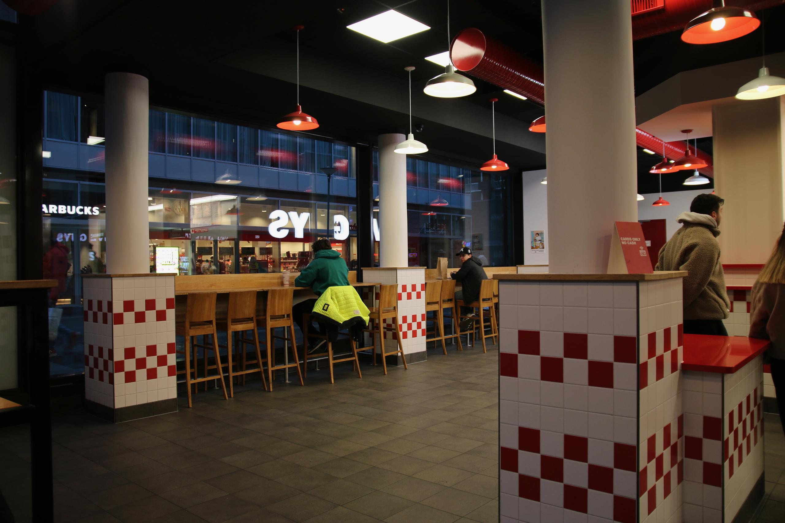 Interior seating area of Five Guys Netherlands with high wooden bar stools, red and white tiles and large windows overlooking the shopping street