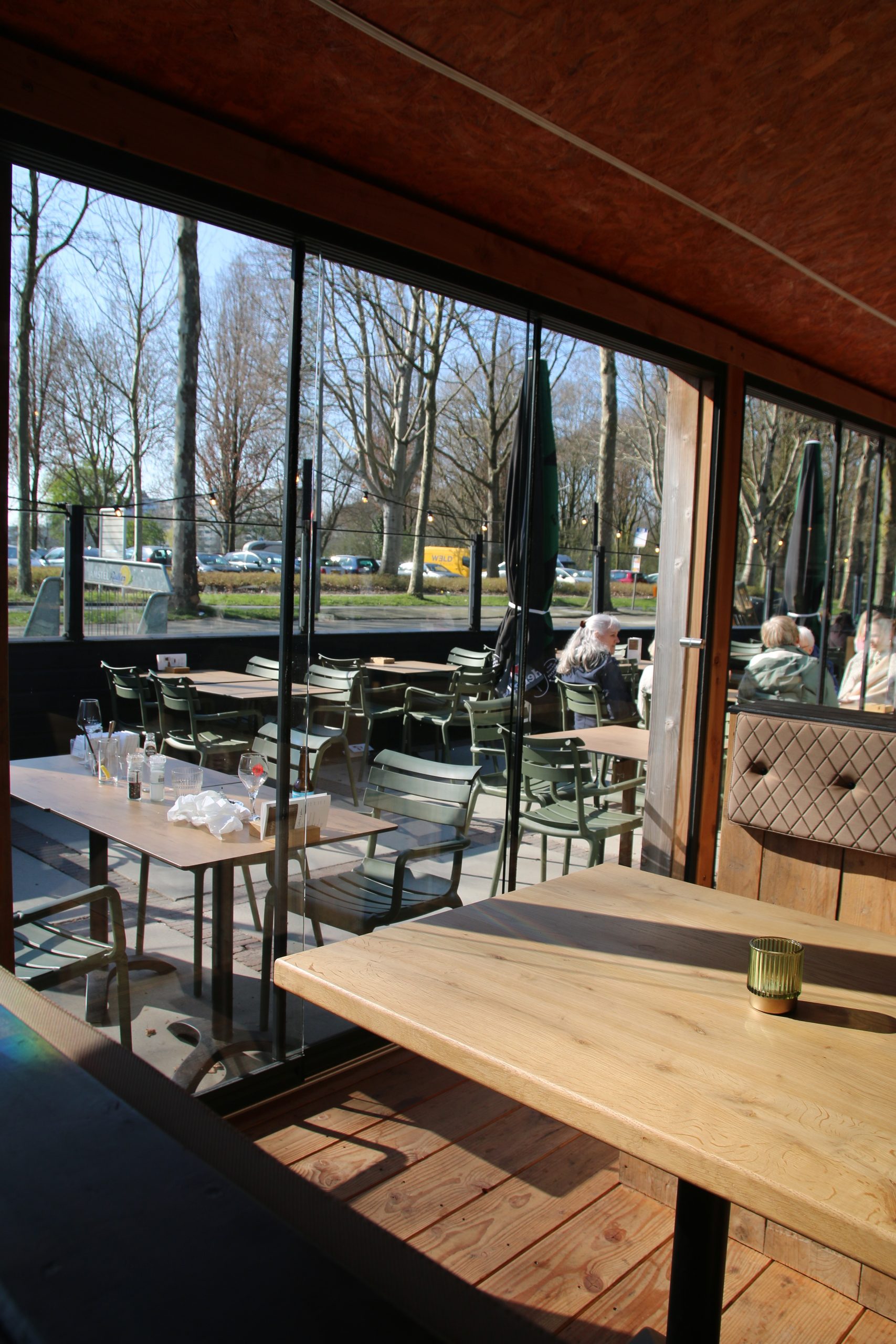 Bij Hen Alphen aan den Rijn cozy covered terrace with wooden tables, glass sliding doors, and sunny seating area