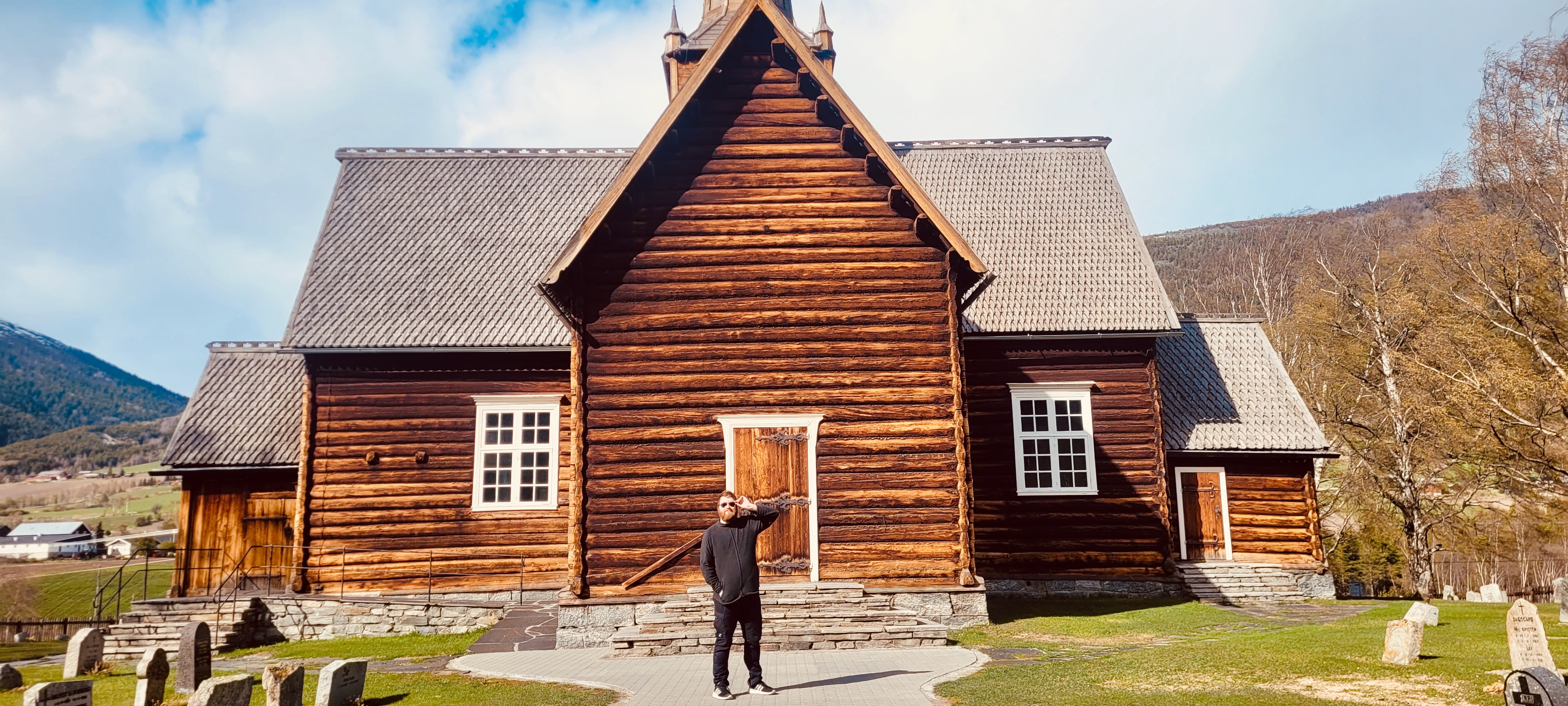 Timo van den Berg standing in front of Lom Stave Church in Lom, Norway, with dark wooden stave architecture, detailed timber walls, stone steps, old gravestones, mountain scenery and a bright cloudy sky.