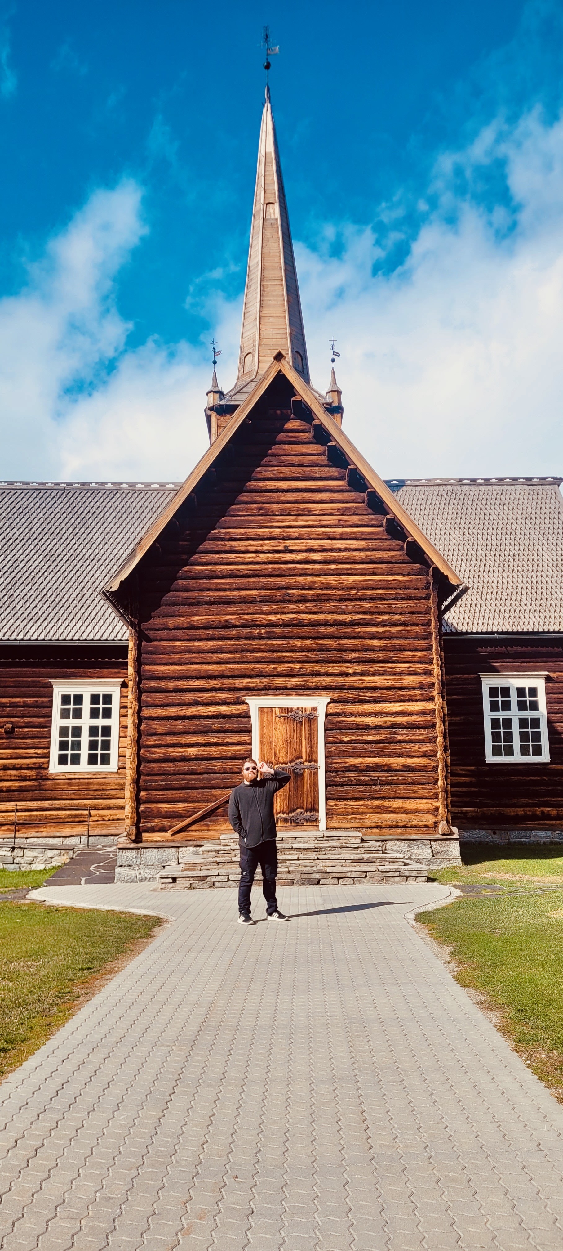 Lom Stave Church in Lom, Norway, with Timo van den Berg standing in front of the wooden entrance, showing the tall central spire, dark timber walls, carved door details and bright blue sky.