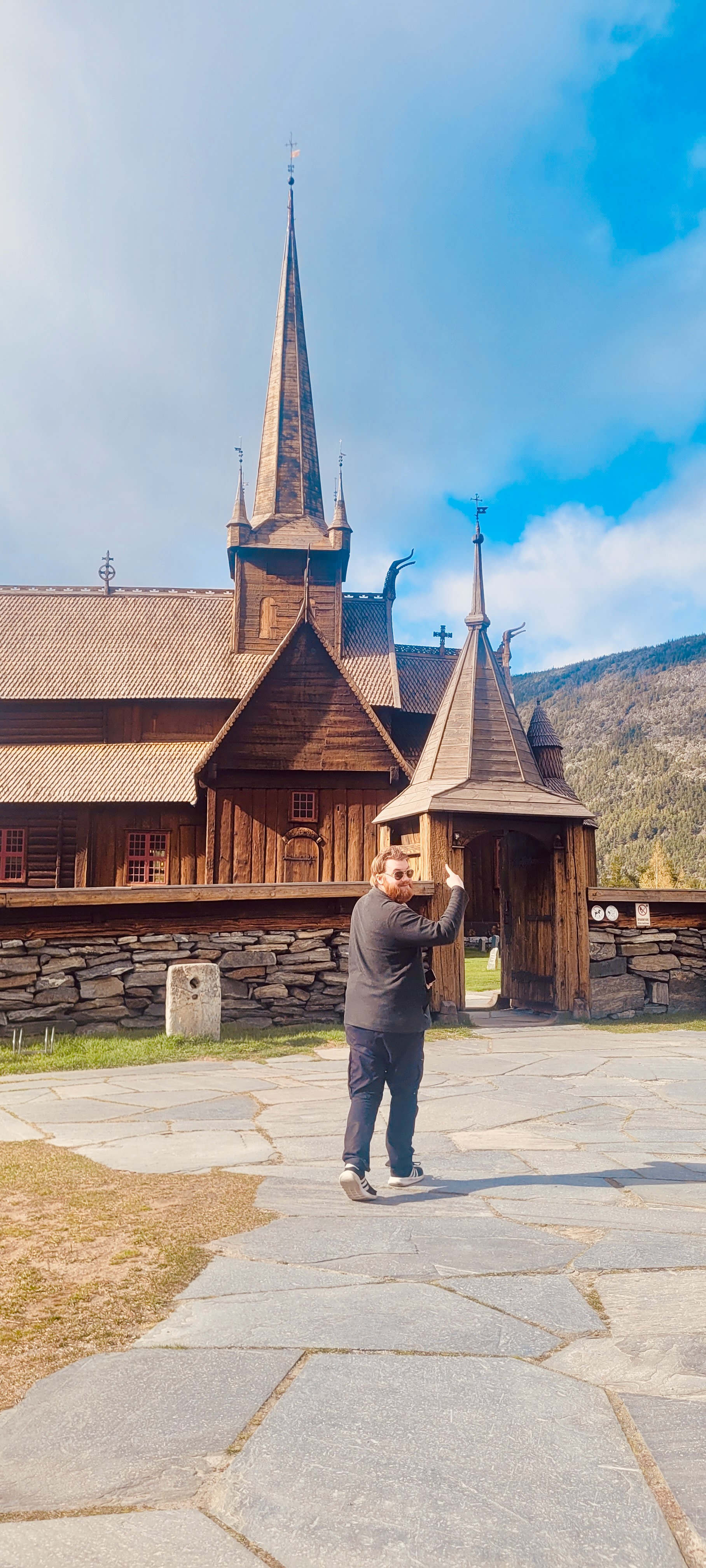 Lom Stave Church in Norway with a visitor, Timo van den Berg, pointing toward the wooden gatehouse, tall church spires, carved roof details, stone wall, mountain background and partly cloudy blue sky.
