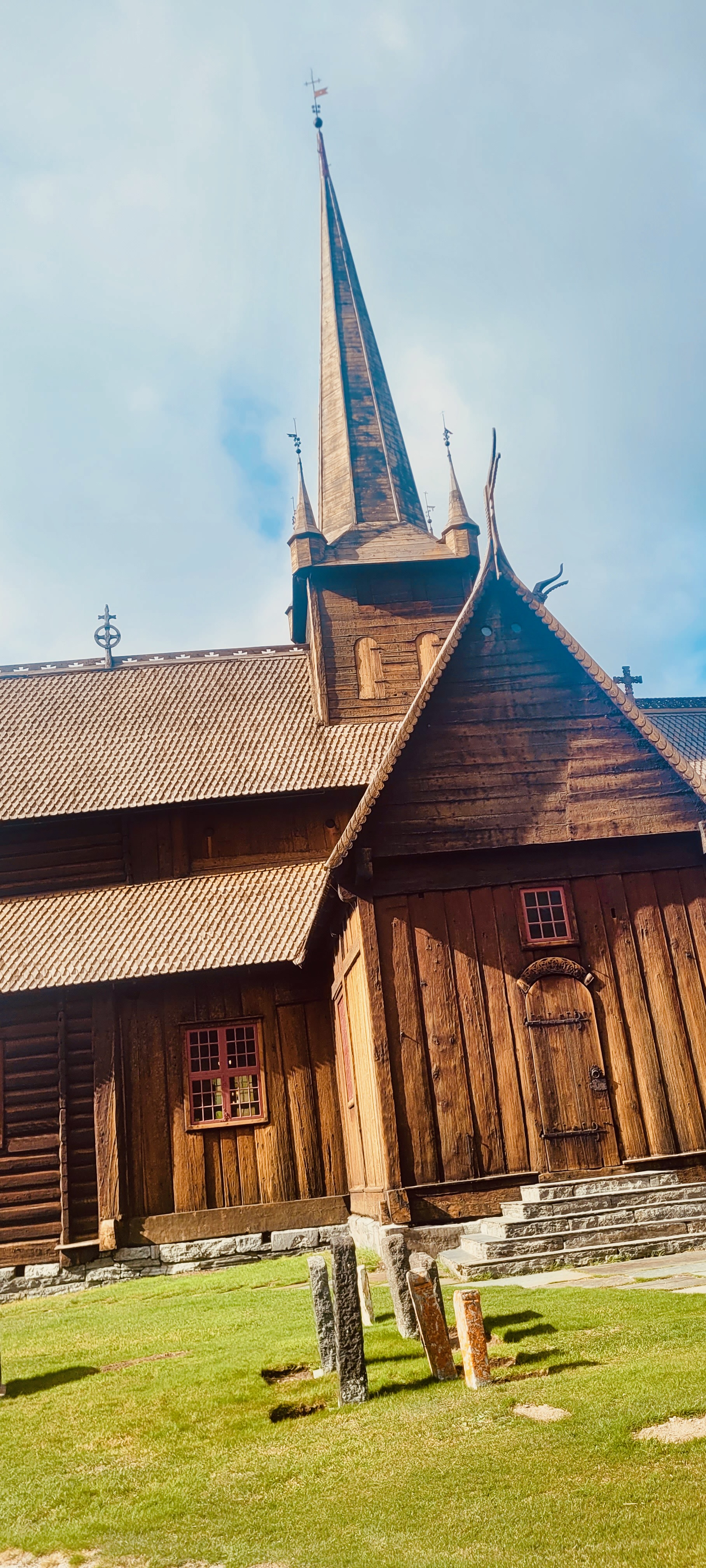 Close view of Lom Stave Church with dark wooden stave architecture, steep rooflines, carved dragon head details, tall spire, small red framed windows and old gravestones in the churchyard.