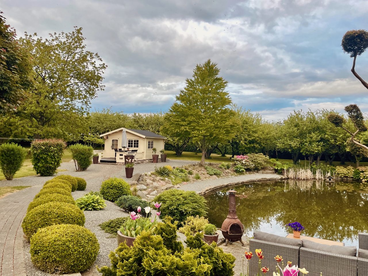Annekset pa Langholm review showing a peaceful wooden Airbnb garden hut beside a pond with flowers trees gravel paths outdoor seating and a cloudy sky in Ringe Denmark