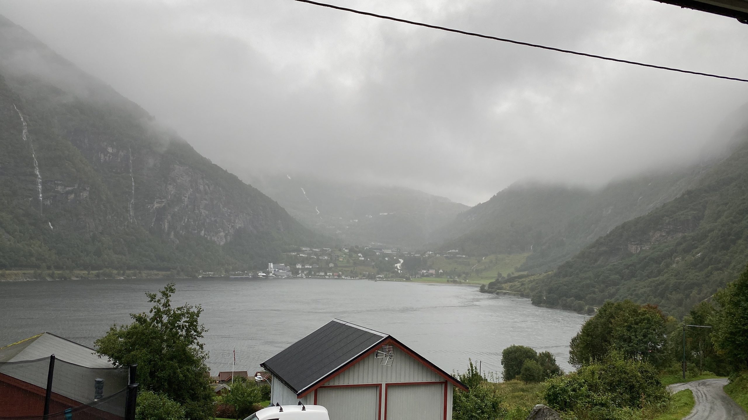 Geiranger Airbnb view over Geirangerfjord with misty mountains, waterfalls, cloudy weather, and the village of Geiranger in the distance