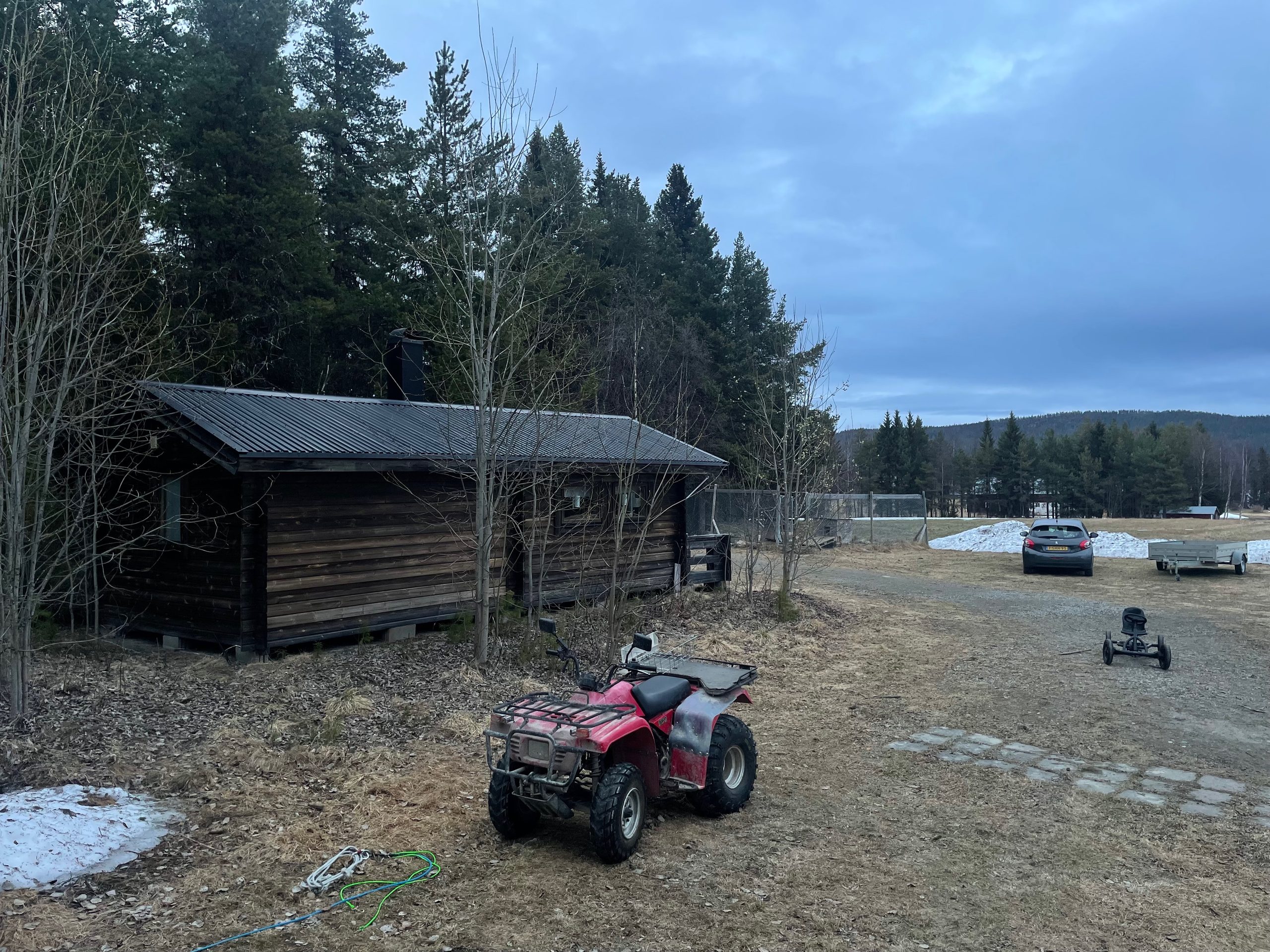 Sorsele Airbnb cabin in Swedish Lapland with a red ATV in the yard, pine forest behind the wooden cabin, patches of melting snow, a gravel driveway, parked car, and quiet countryside surroundings in early spring.