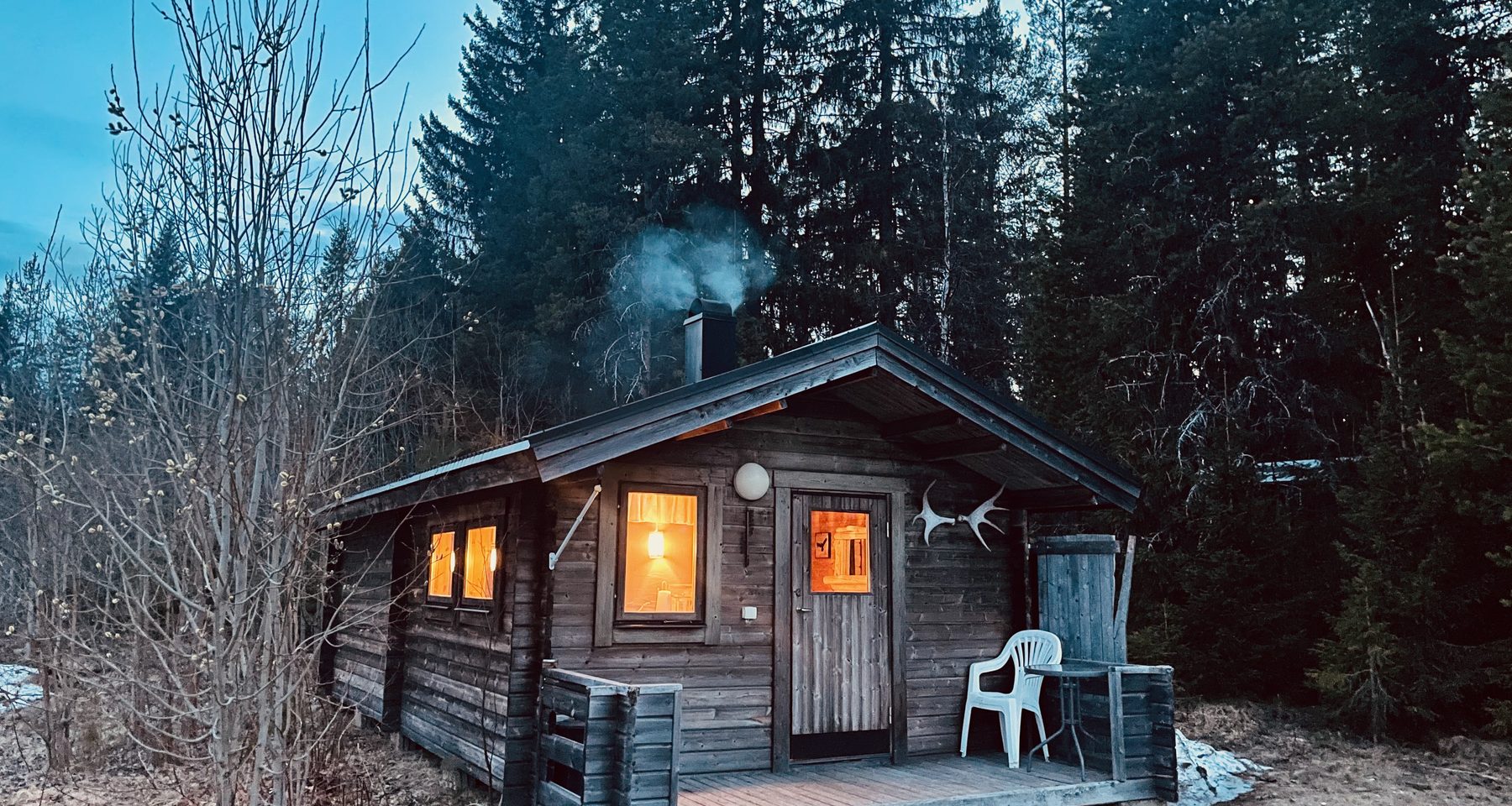 Sorsele Airbnb cabin in Swedish Lapland at dusk, with warm lights glowing through the windows, smoke rising from the fireplace chimney, a small wooden porch with a chair, and dark pine forest surrounding the cozy cabin.