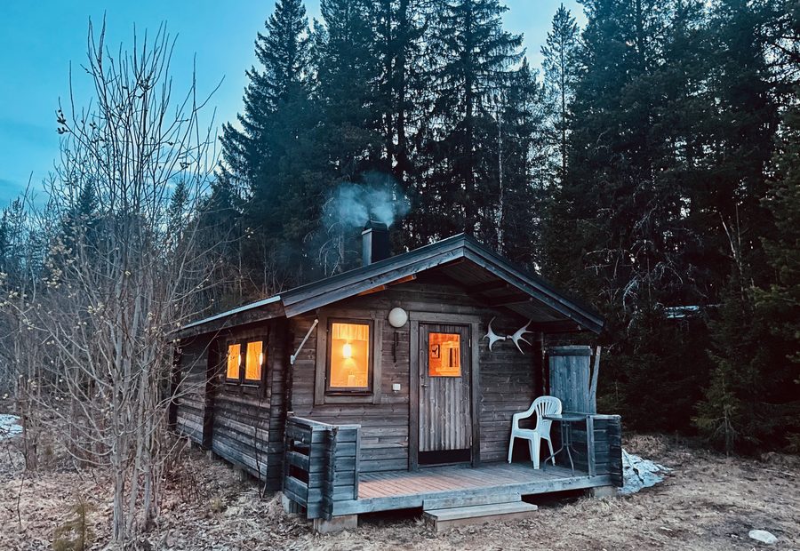 Sorsele Airbnb cabin in Swedish Lapland at dusk, with warm lights glowing through the windows, smoke rising from the fireplace chimney, a small wooden porch with a chair, and dark pine forest surrounding the cozy cabin.