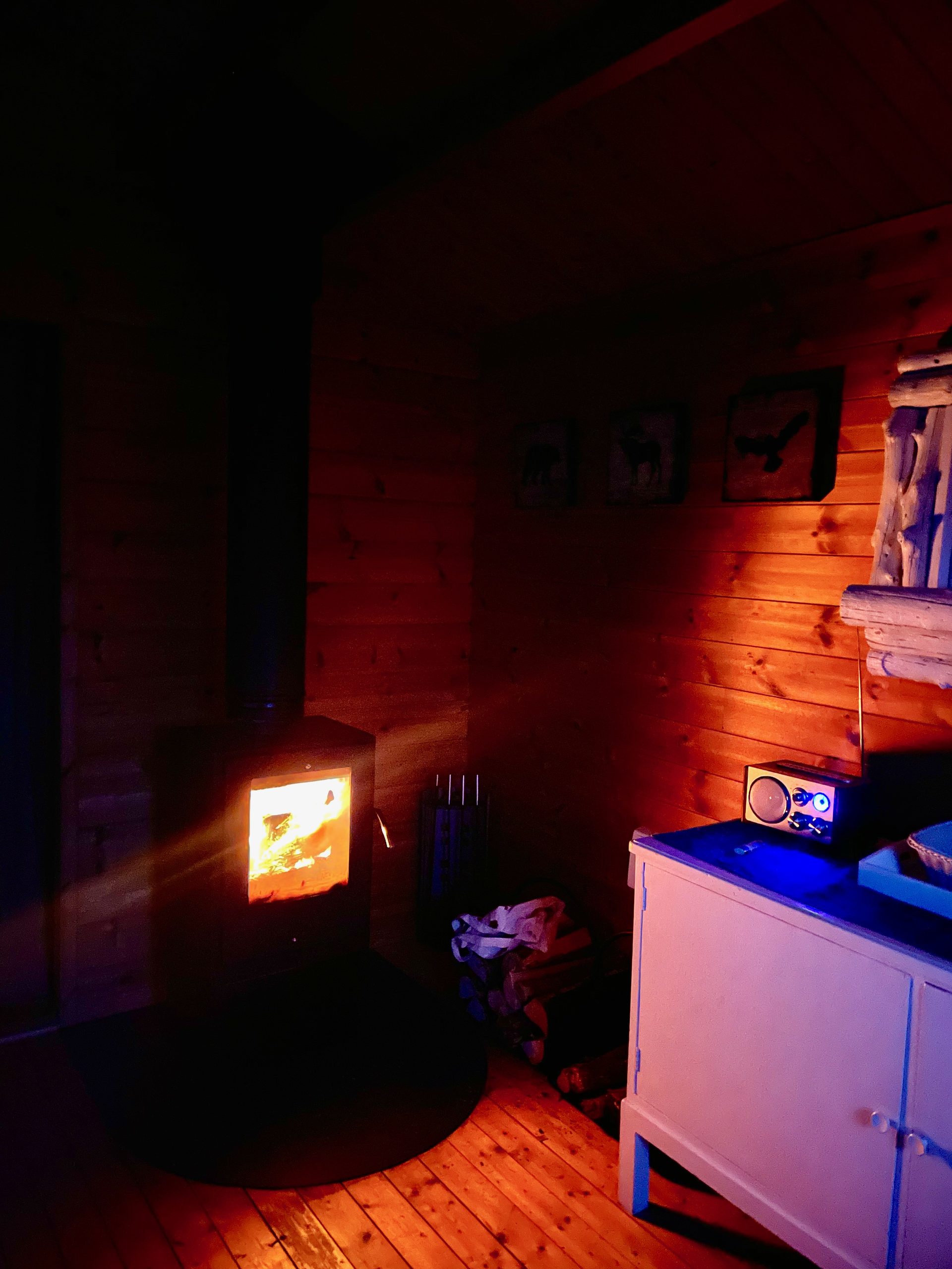 Sorsele Airbnb cabin at night with the wood burning stove glowing warmly, red and orange firelight reflecting on the wooden walls, an FM radio on the cabinet, and a cozy Lapland cabin atmosphere.