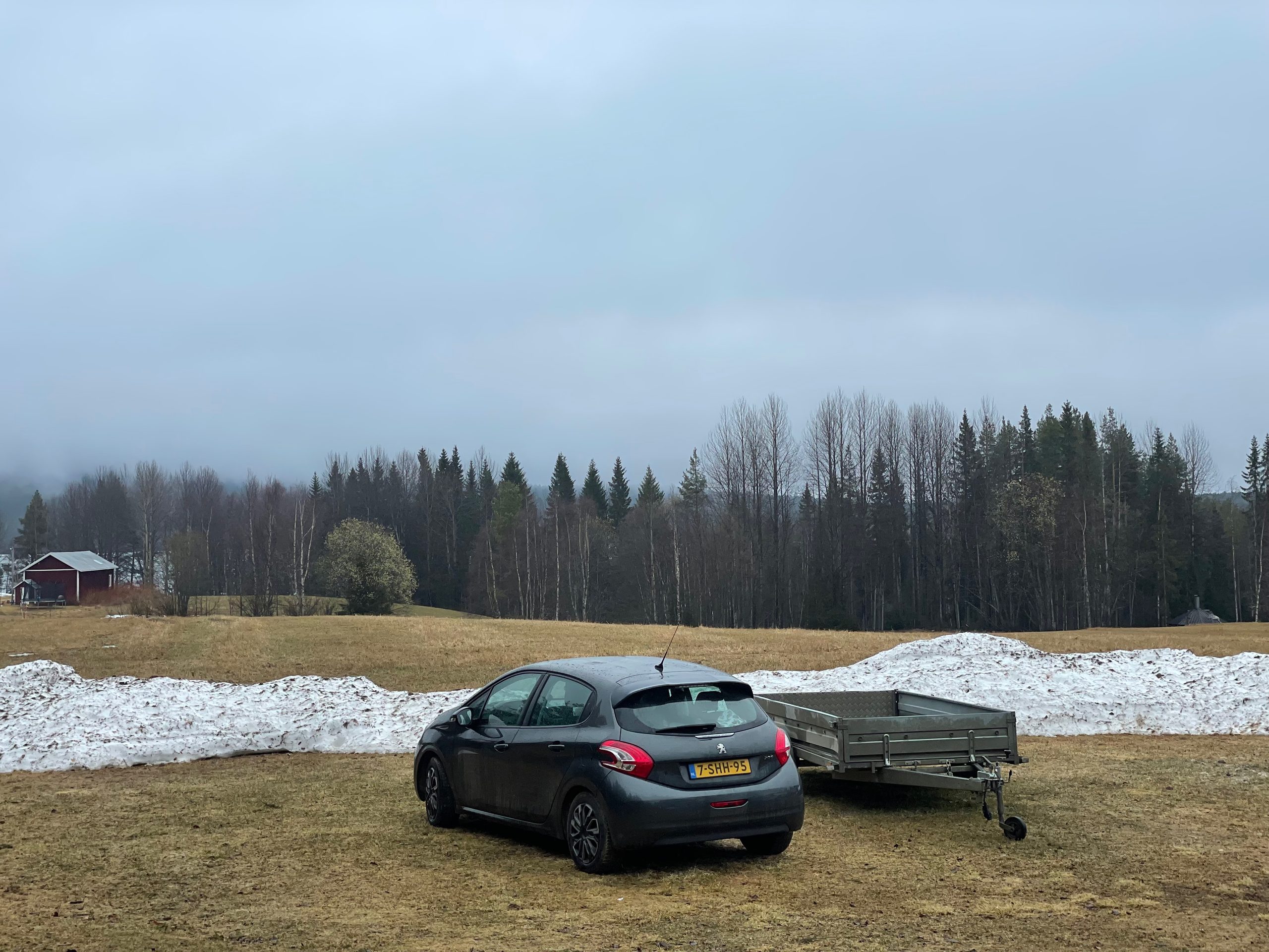 Sorsele Airbnb surroundings in Swedish Lapland with the Timobiel, a Peugeot 208, parked beside a trailer near melting snow, open fields, dark forest, and a quiet rural Lapland landscape in early spring.