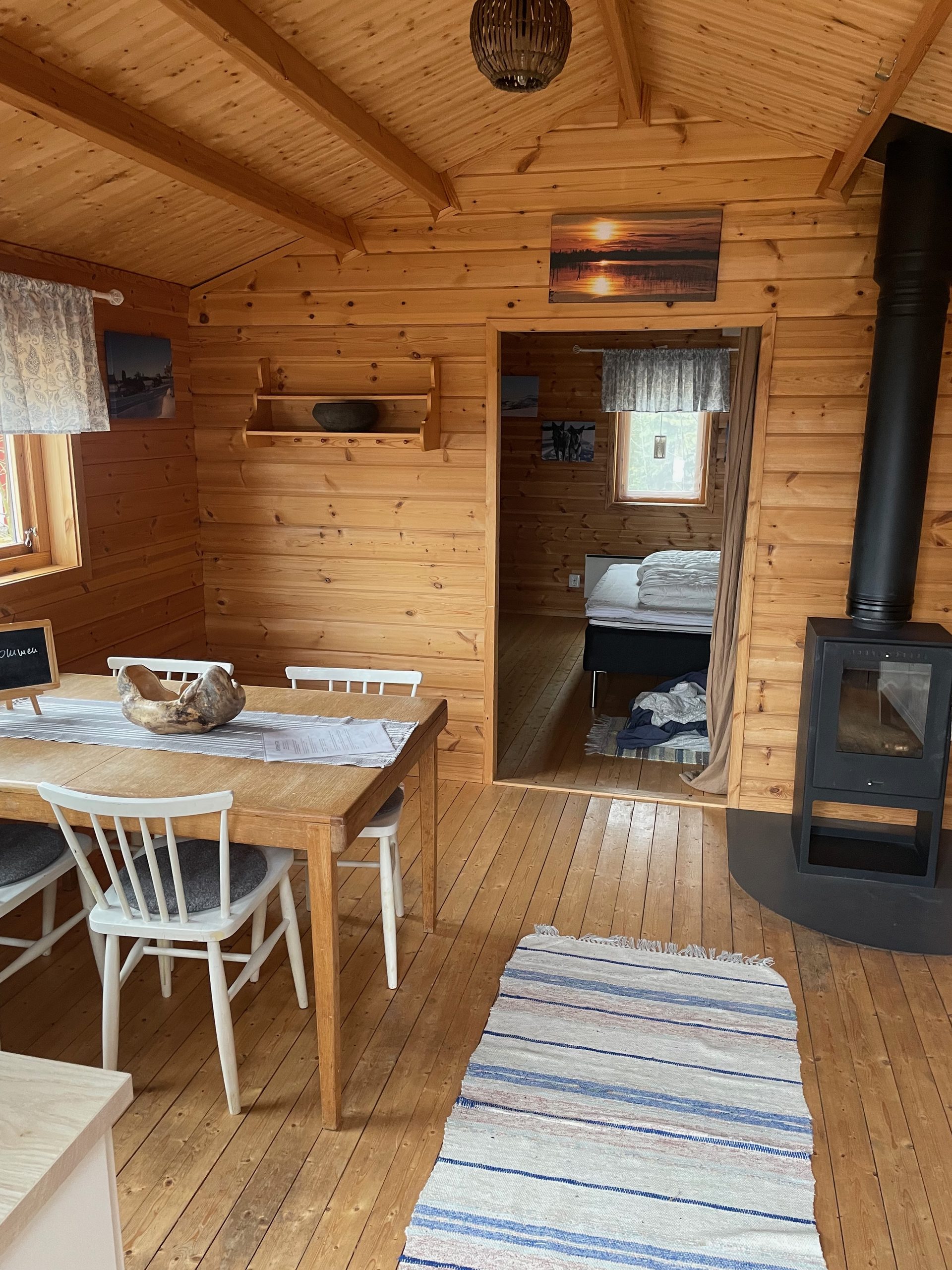 Sorsele Airbnb interior with a bright wooden cabin living area, dining table, white chairs, striped rug, wood burning stove, open doorway to the bedroom, and a cozy double bed visible in the back room.
