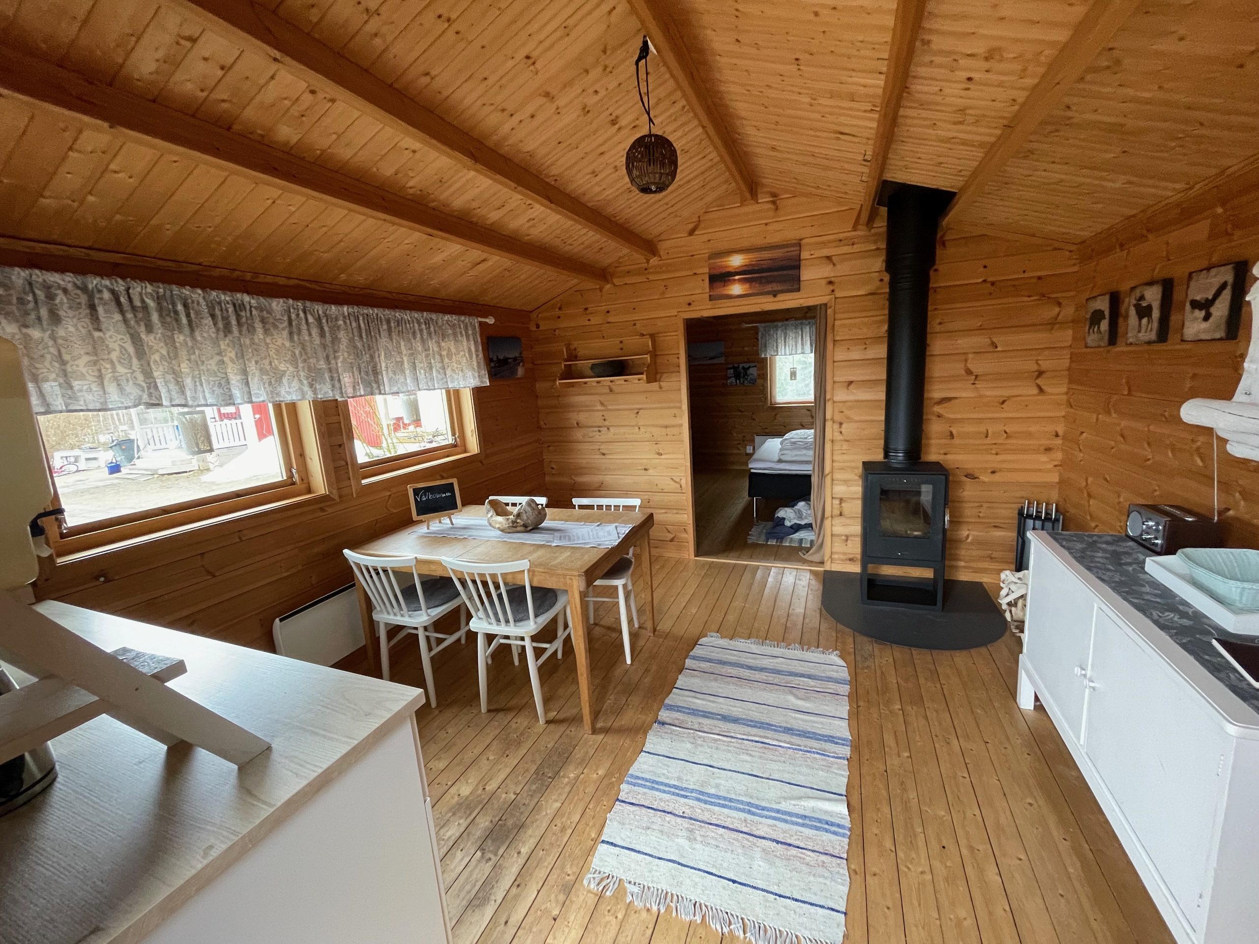 Sorsele Airbnb interior with a spacious wooden living area, dining table with white chairs, striped rug, wood burning stove, small kitchenette, FM radio, and an open doorway leading into the bedroom.