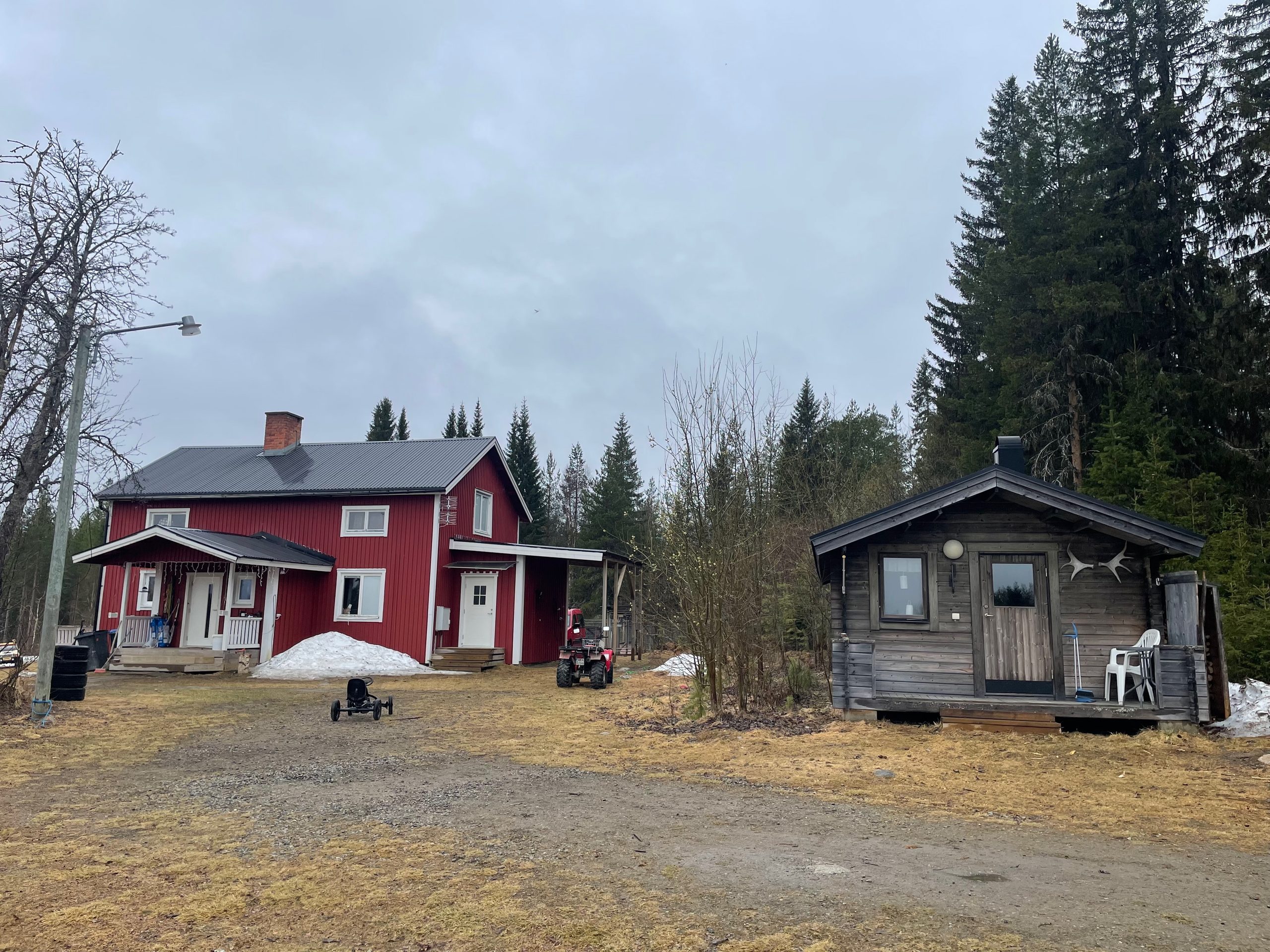 Sorsele Airbnb property in Swedish Lapland with the cozy wooden guest cabin beside a traditional red Swedish house, melting snow on the grass, an ATV near the house, and dark forest surrounding the rural yard.