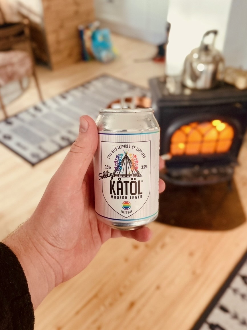Northern Sweden Airbnb review, hand holding a can of Kåta Modern Lager beer in front of a glowing fireplace inside a cozy Swedish Airbnb cabin