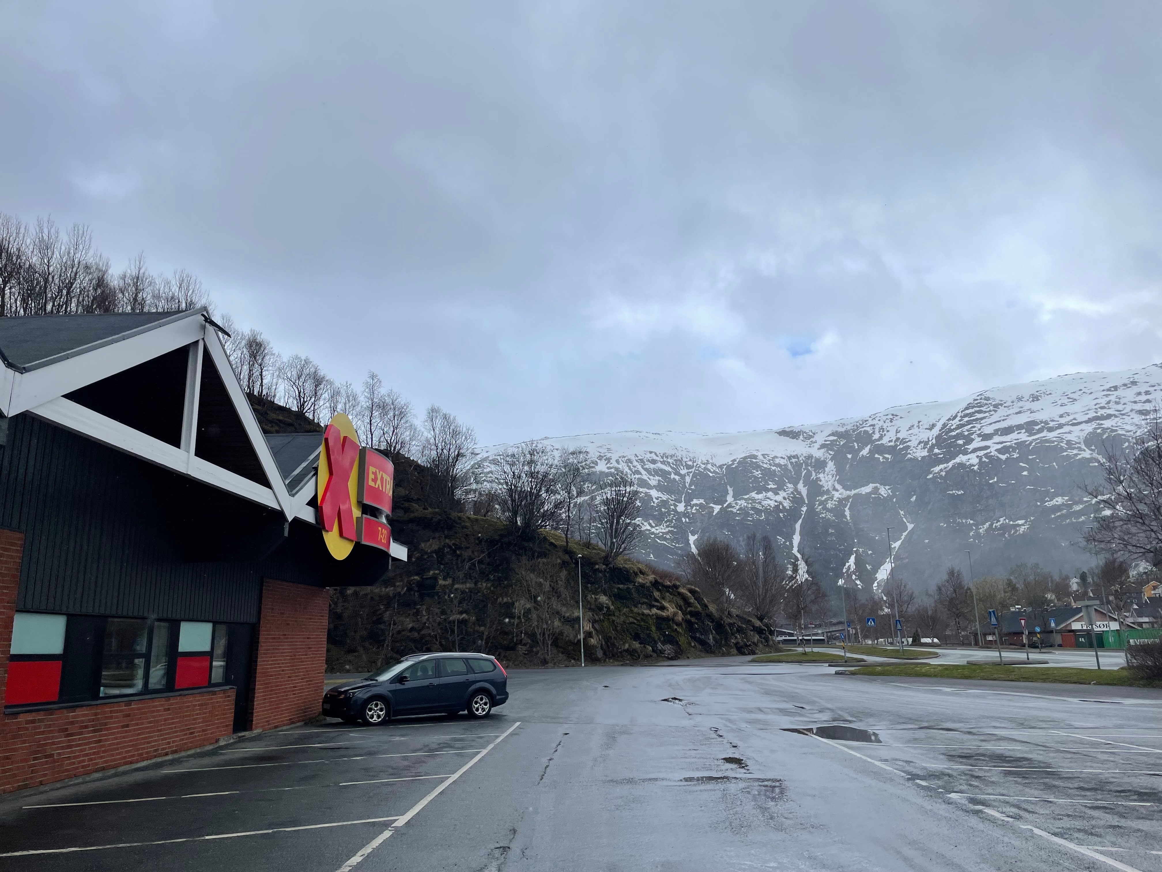 Extra Halsøy supermarket parking lot in Mosjøen, Norway, with the store building, Extra sign, wet road surface, parked car, and snow-covered mountains behind it.