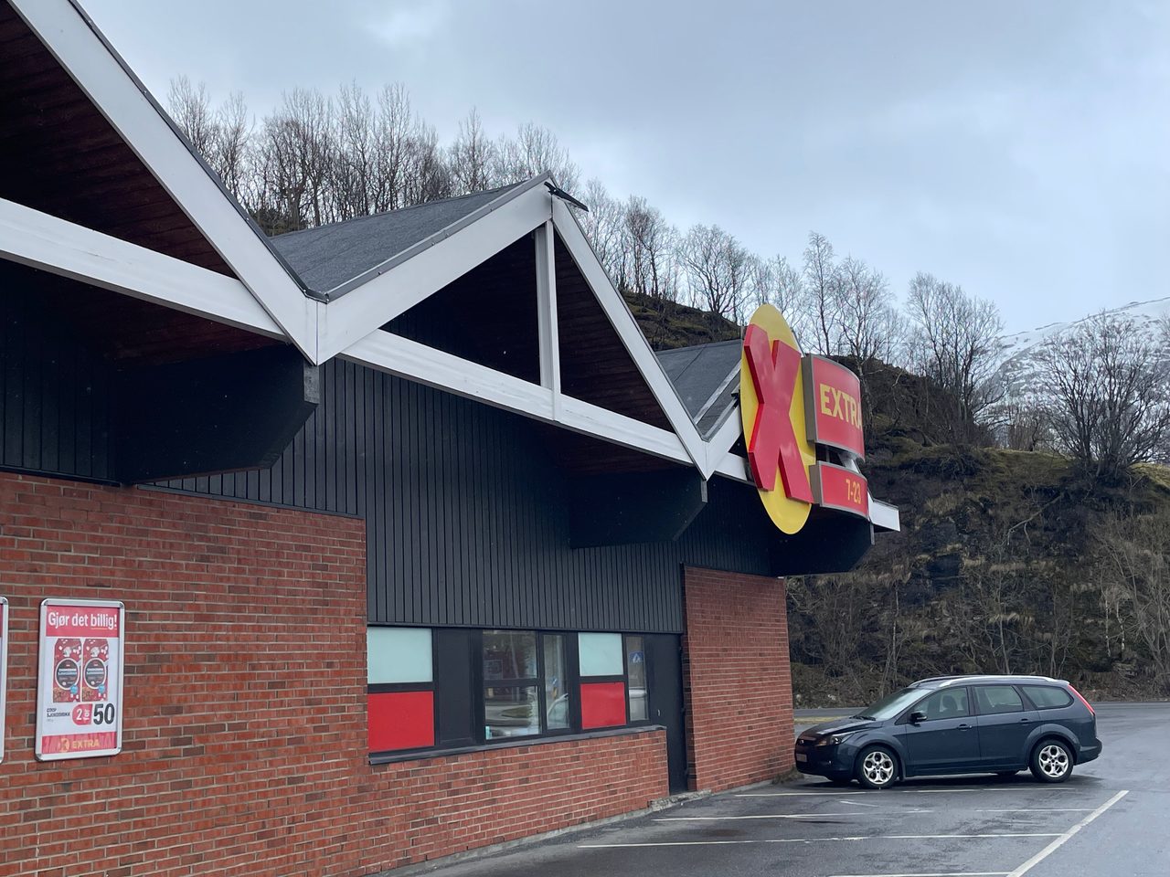 Extra Halsøy supermarket exterior in Mosjøen, Norway, with the red and yellow Extra sign, brick building, parking area, and snowy mountains in the background.