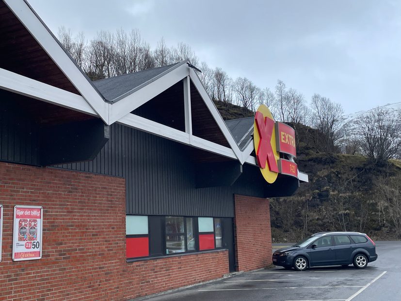 Extra Halsøy supermarket exterior in Mosjøen, Norway, with the red and yellow Extra sign, brick building, parking area, and snowy mountains in the background.